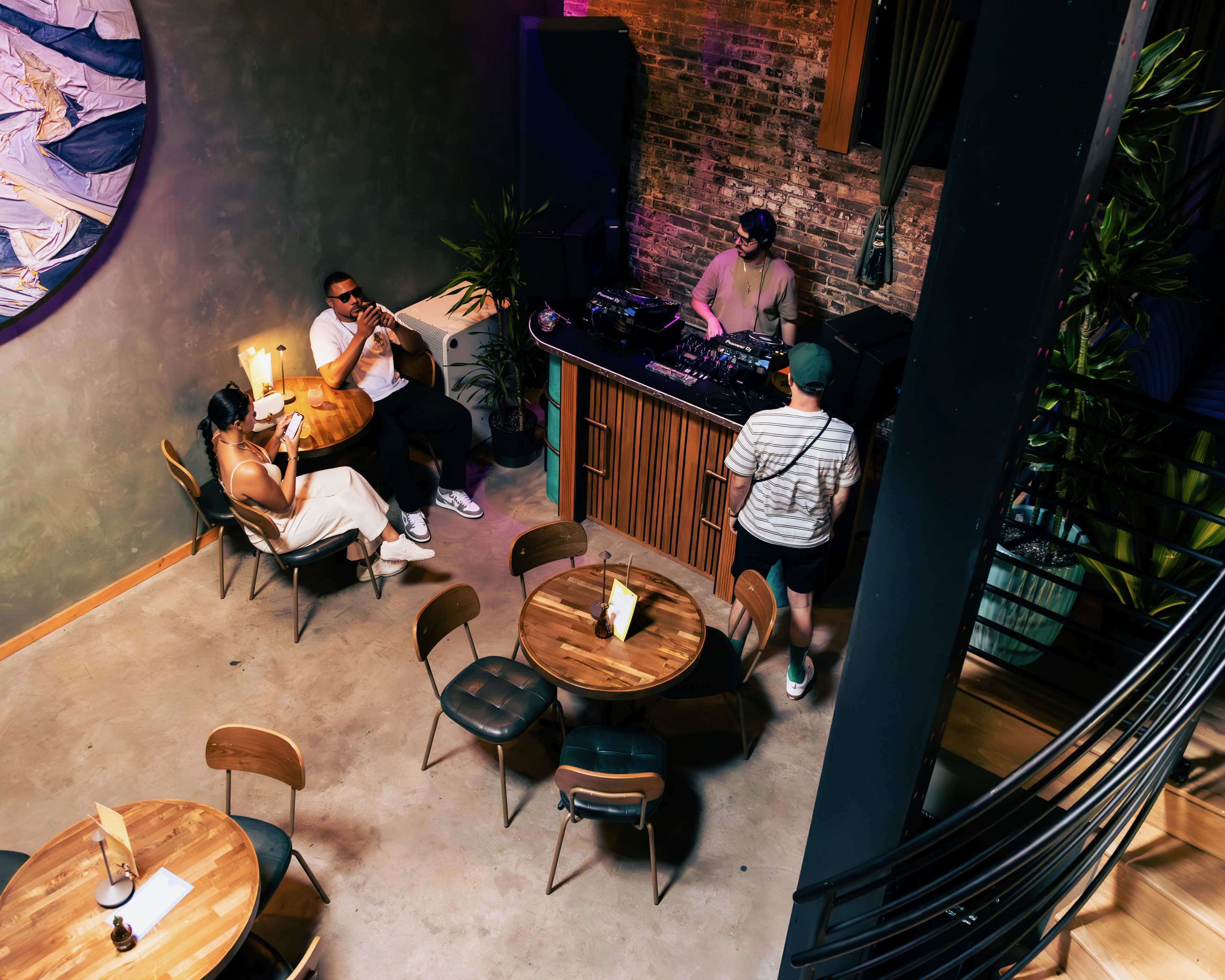 A DJ performs at the front of a stylish restaurant while patrons sit at wooden tables.