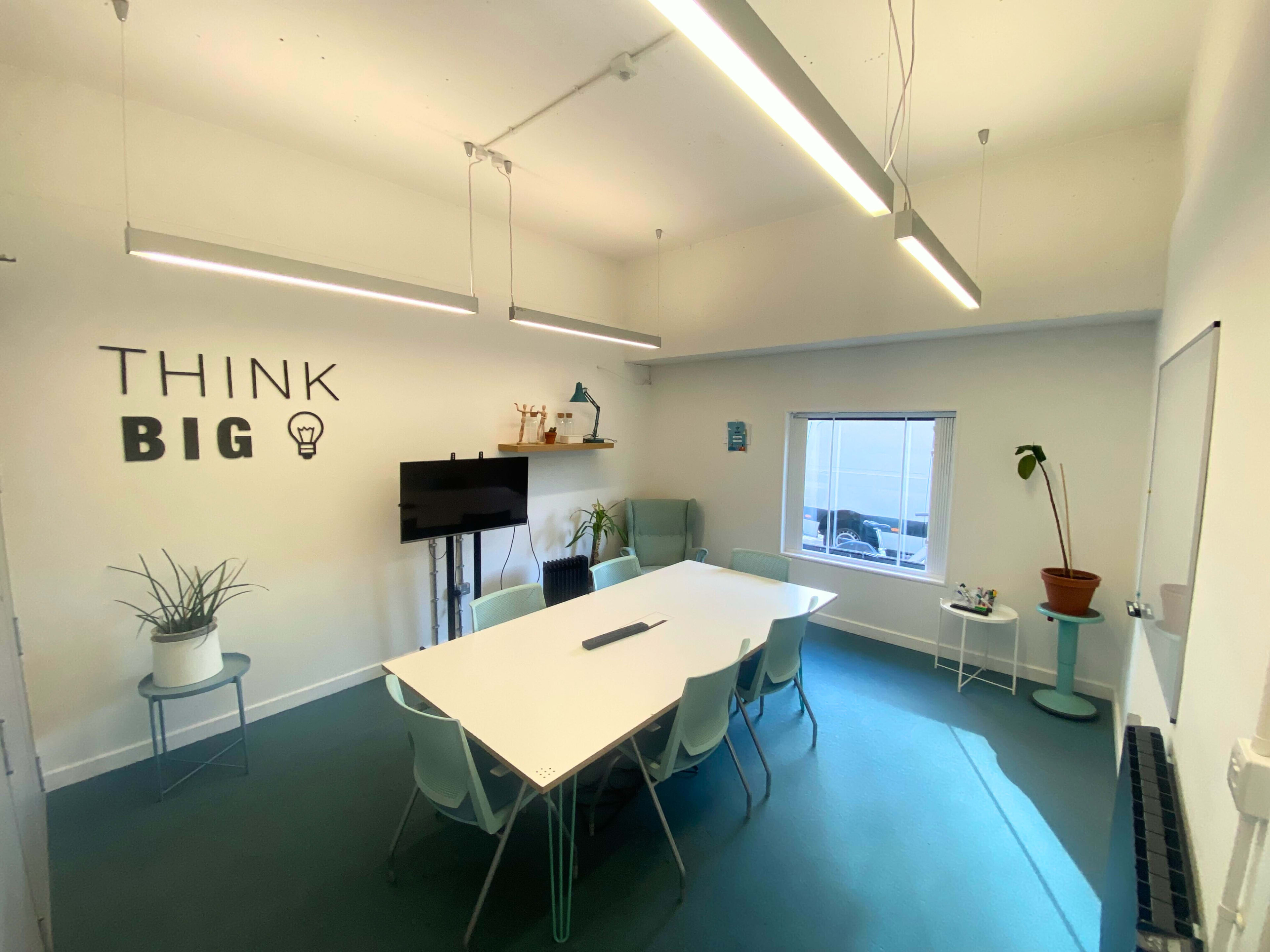 A well-lit meeting room with a large white table surrounded by light-colored chairs, a television on the wall, and the words "THINK BIG" displayed decoratively.