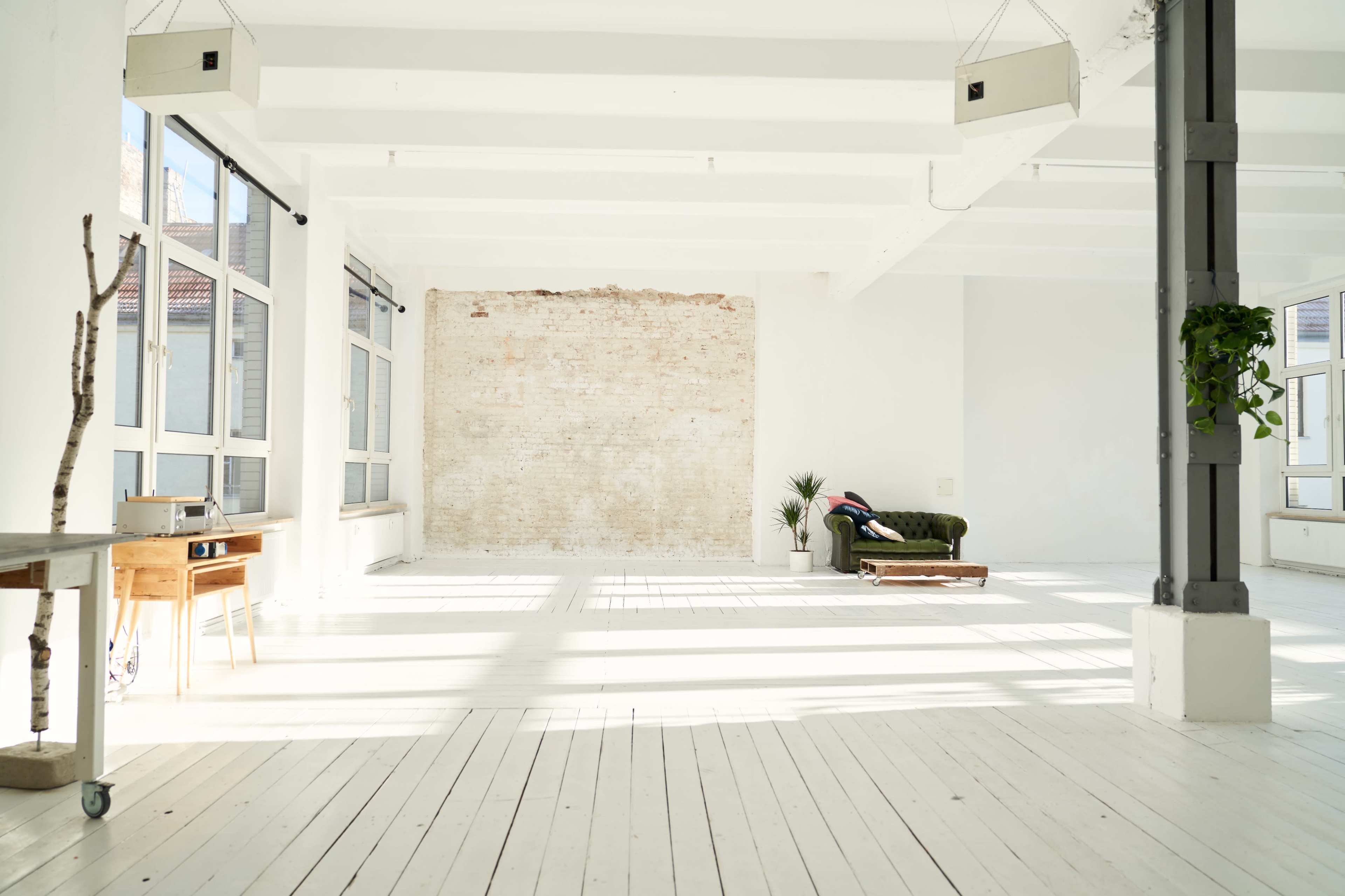 A bright, spacious room with white walls, large windows, and a wooden floor, featuring a sofa and a small table.