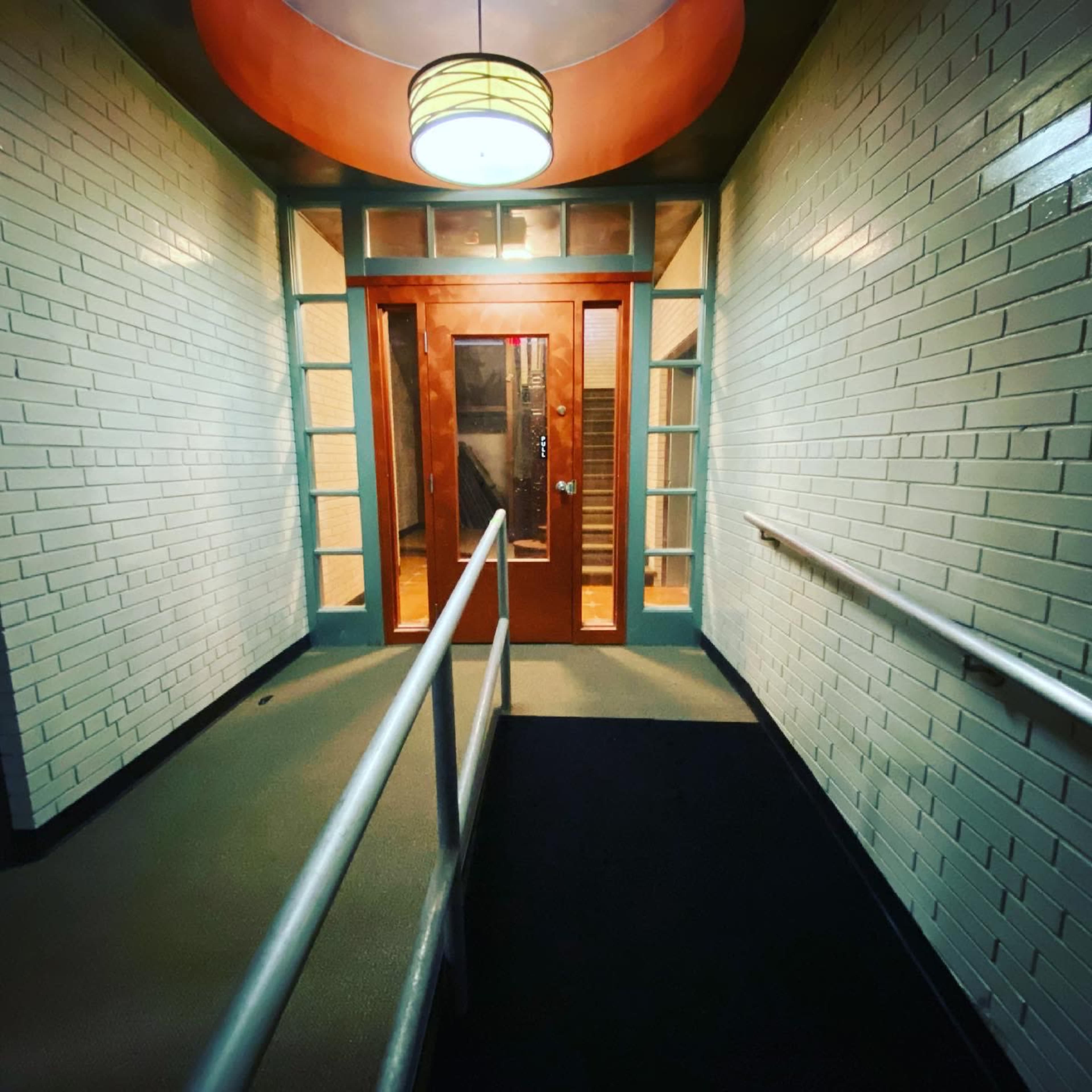 A hallway leading to a wooden door, with white tiled walls and a ceiling light fixture above.