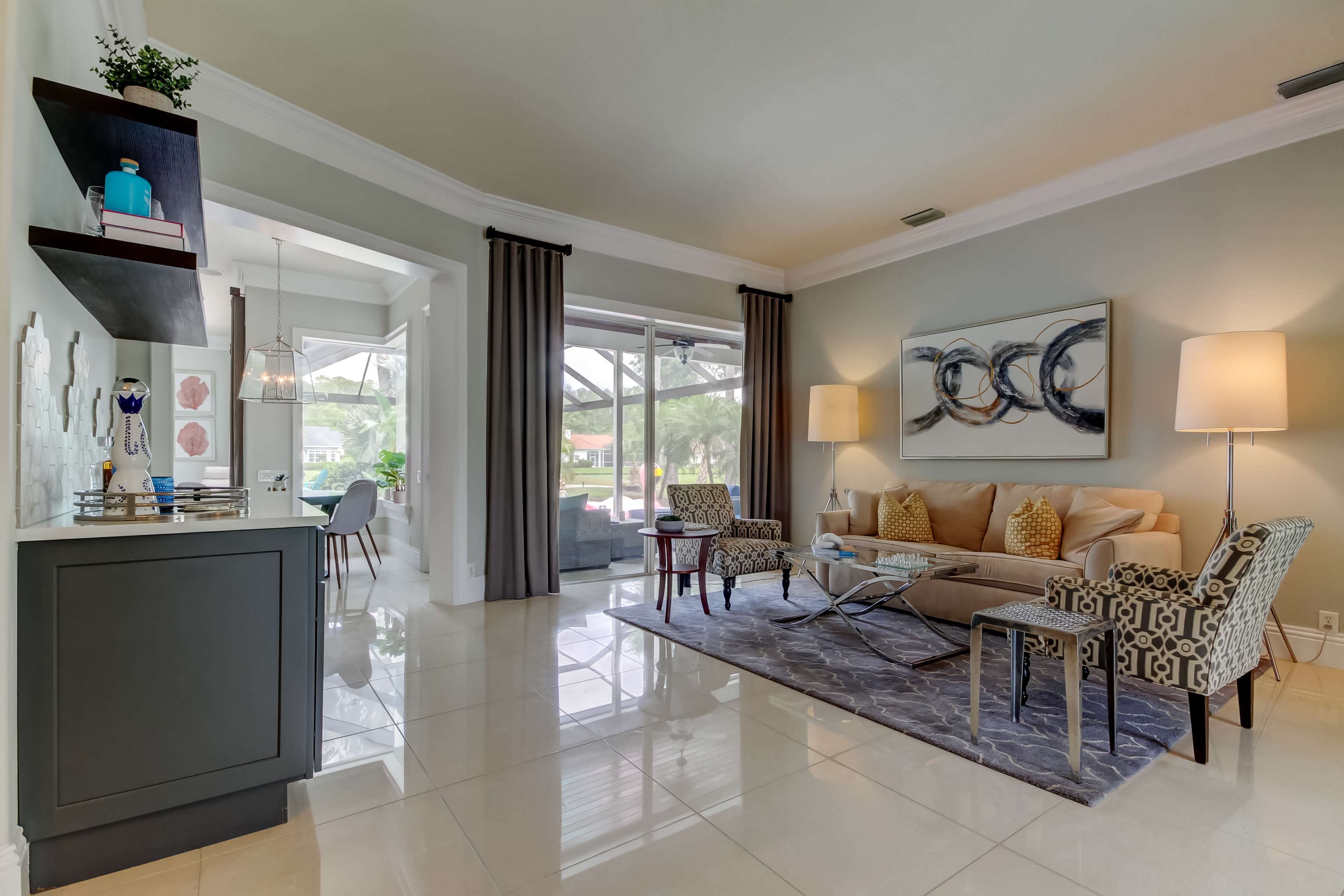 The image shows a modern living room with a beige sofa, patterned chairs, a glass coffee table, and large windows letting in natural light.