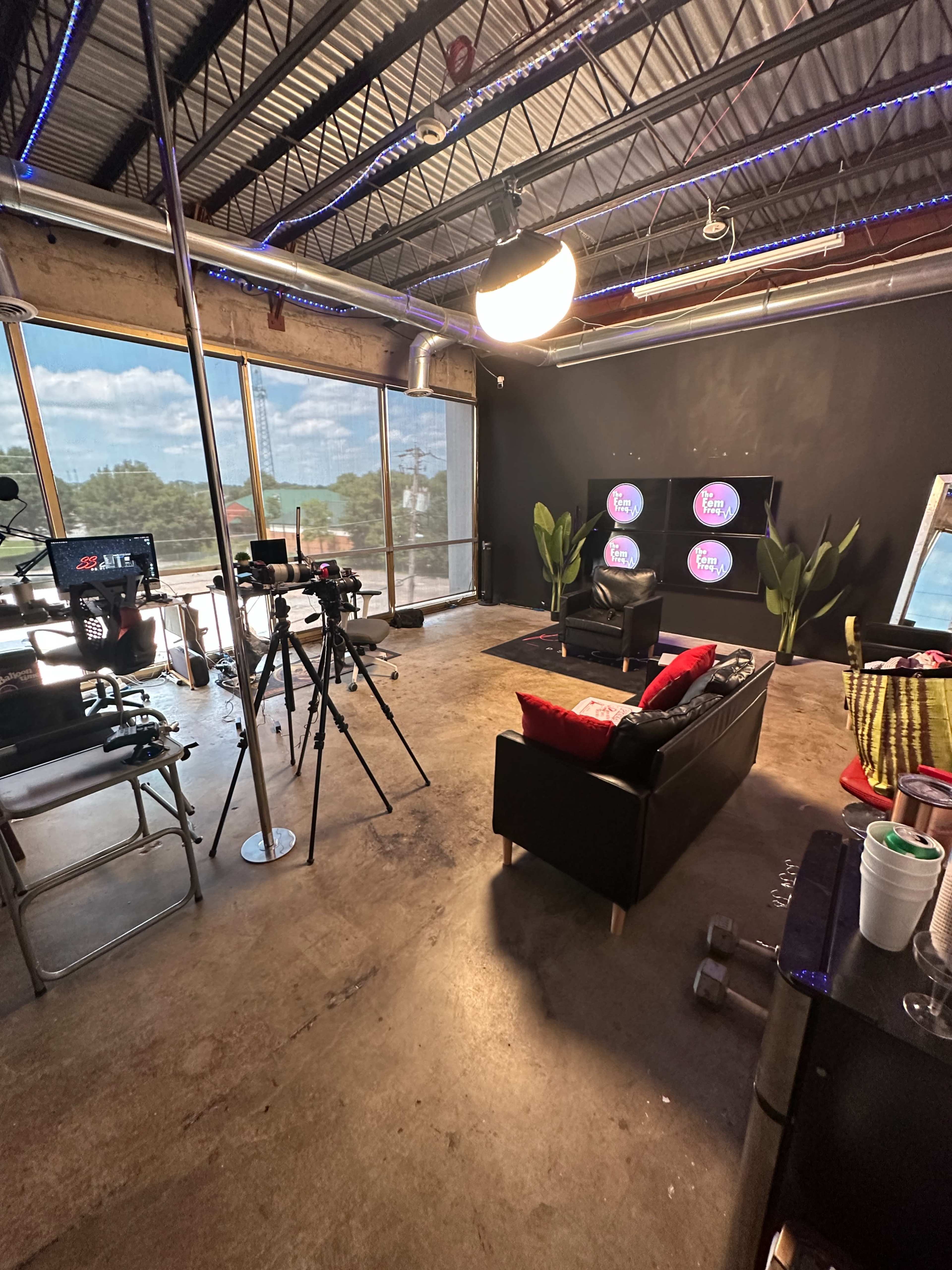 A modern studio setup with a black wall, a couch with red cushions, cameras on tripods, and large screens displaying logos, all under natural light from large windows.