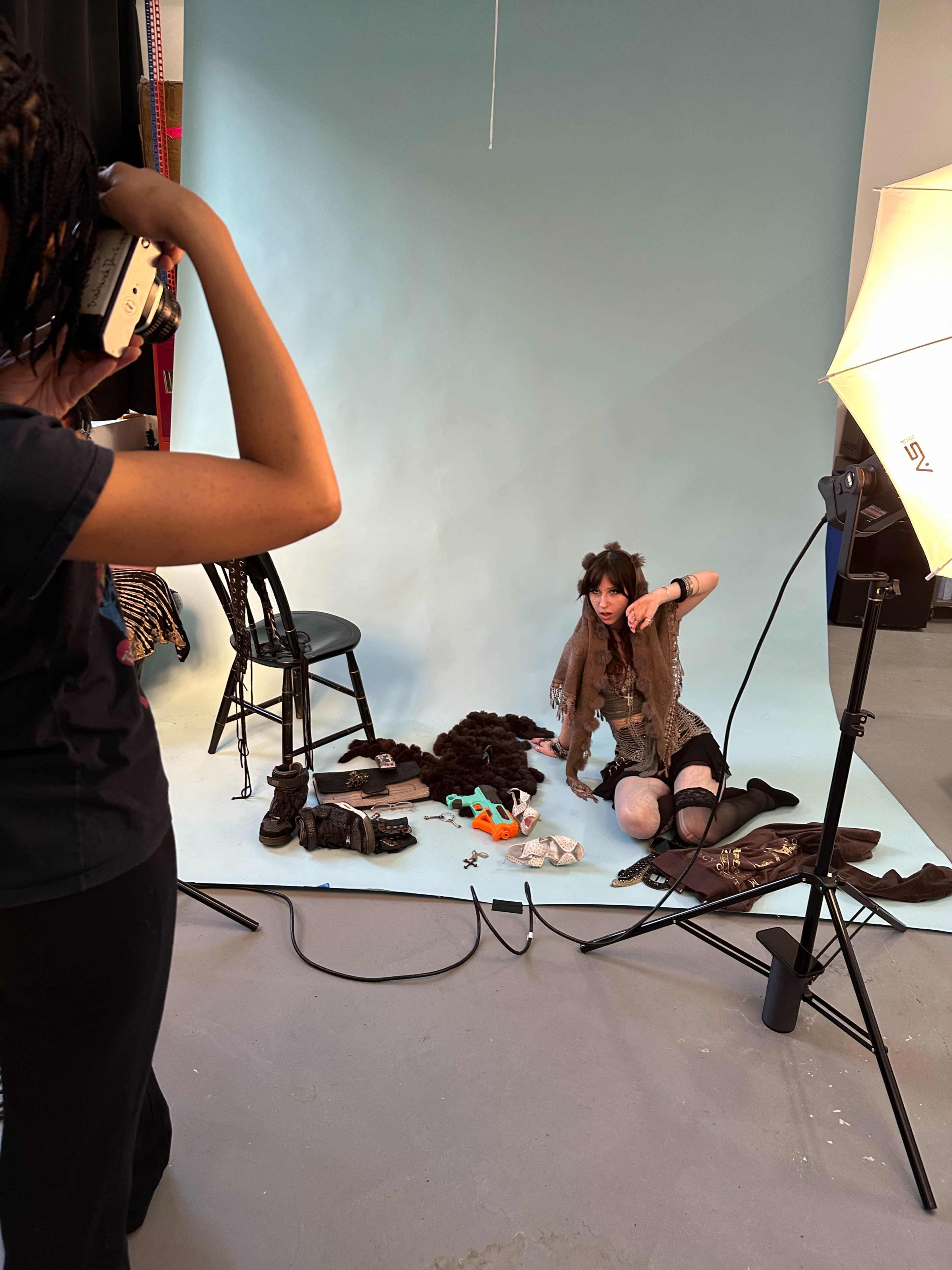 A photographer captures a model posing on the floor in front of a light blue backdrop, surrounded by various props and clothing items.