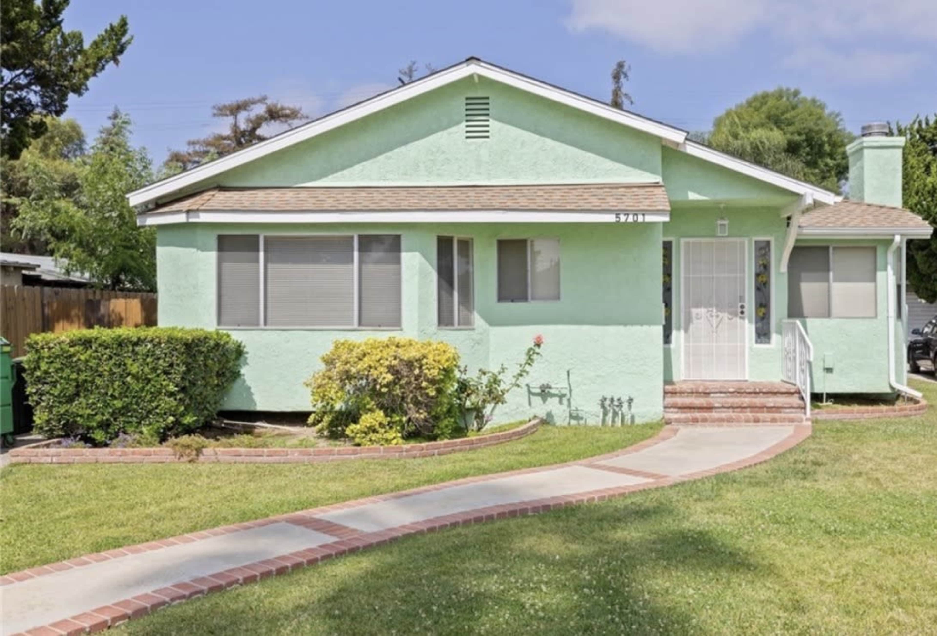 The image shows a light green single-story house with a front yard featuring a walkway and manicured bushes.