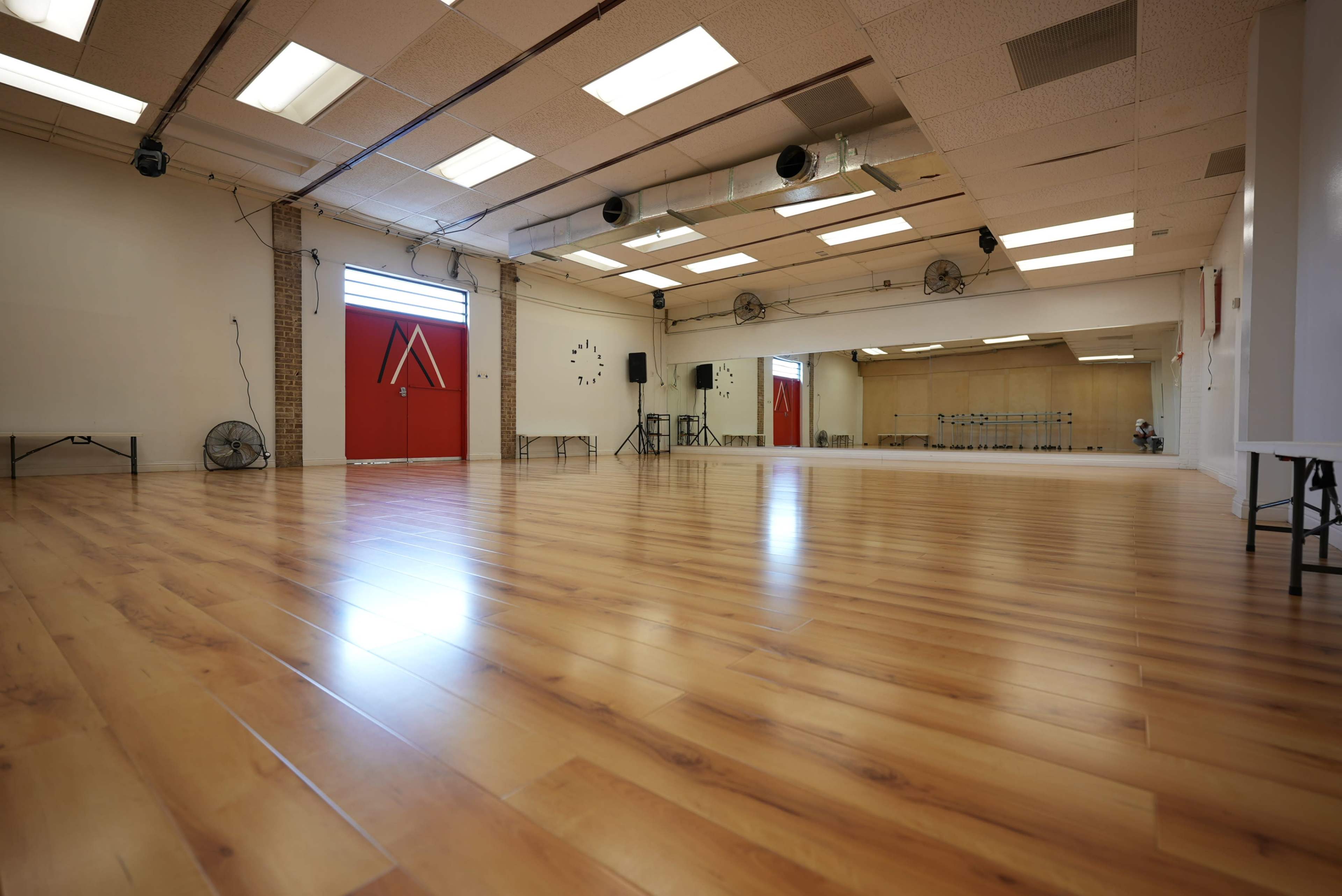 The image shows a large, empty dance studio with hardwood floors and wall-mounted mirrors, featuring red doors at one end.