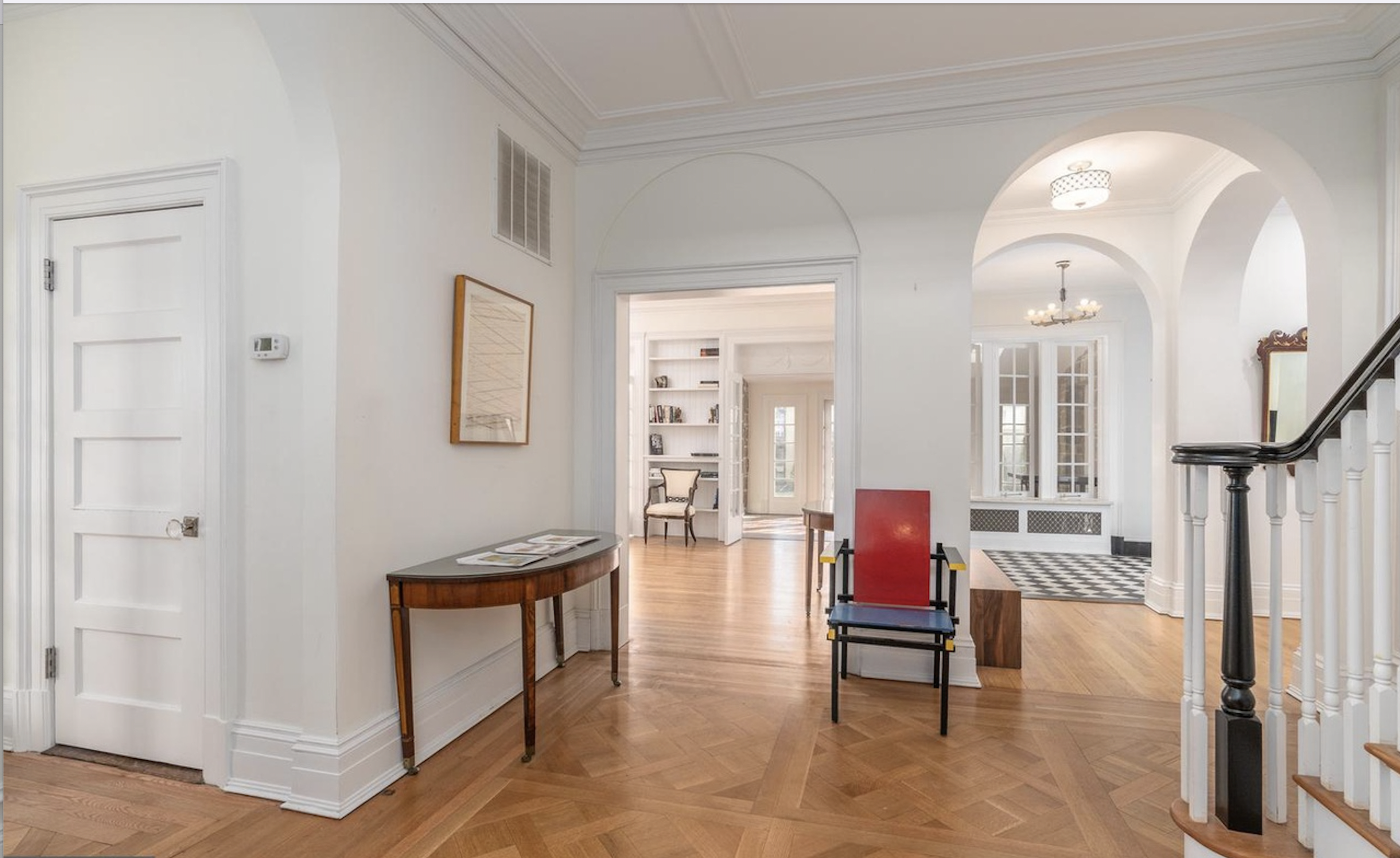 A well-lit hallway with a wooden table and a red chair, leading to another room with arched doorways and a checkered floor.