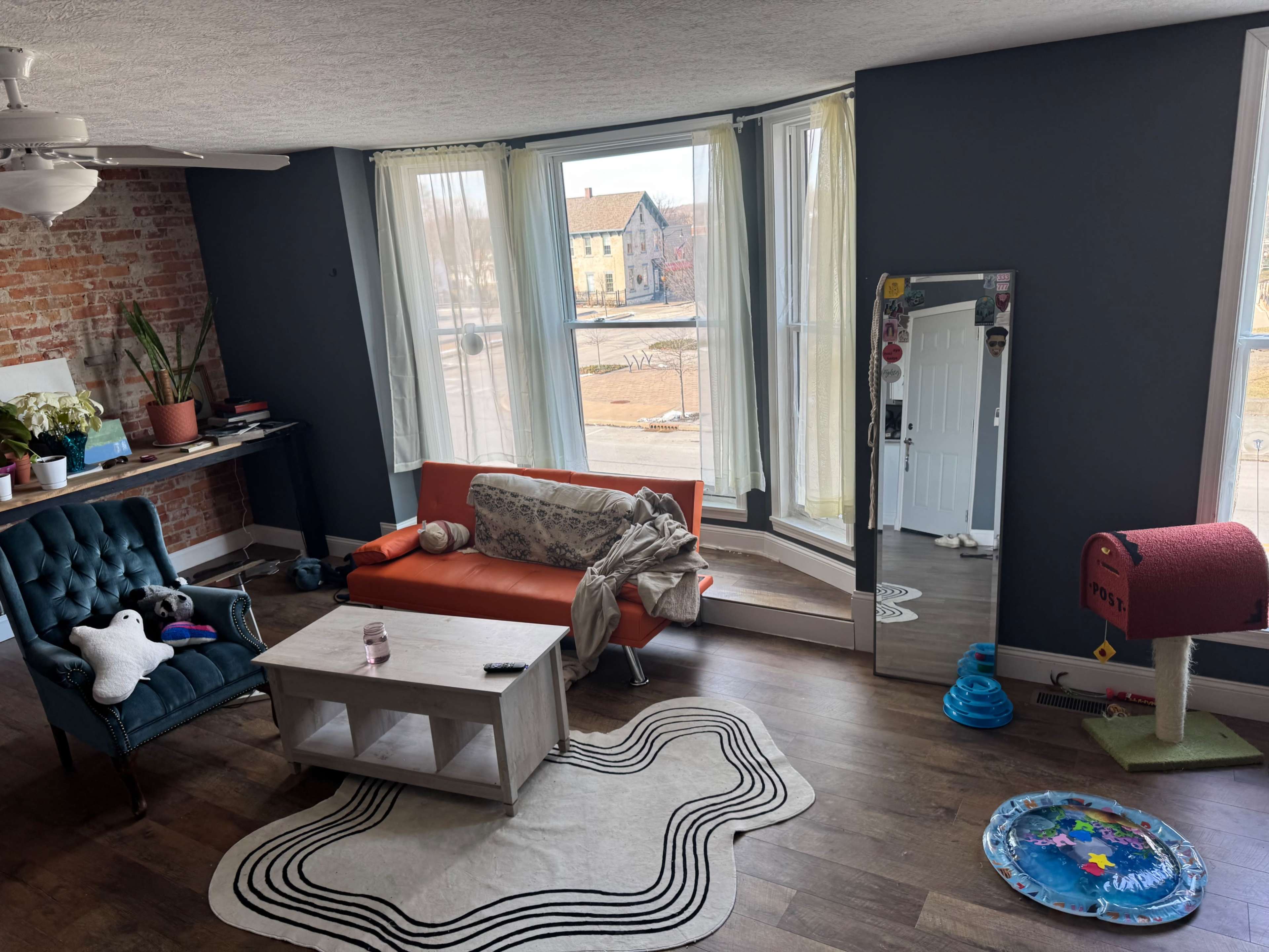 The image shows a living room with a blue wall, a brick accent wall, a green armchair, an orange sofa, a coffee table, and a large mirror next to a window with sheer curtains.