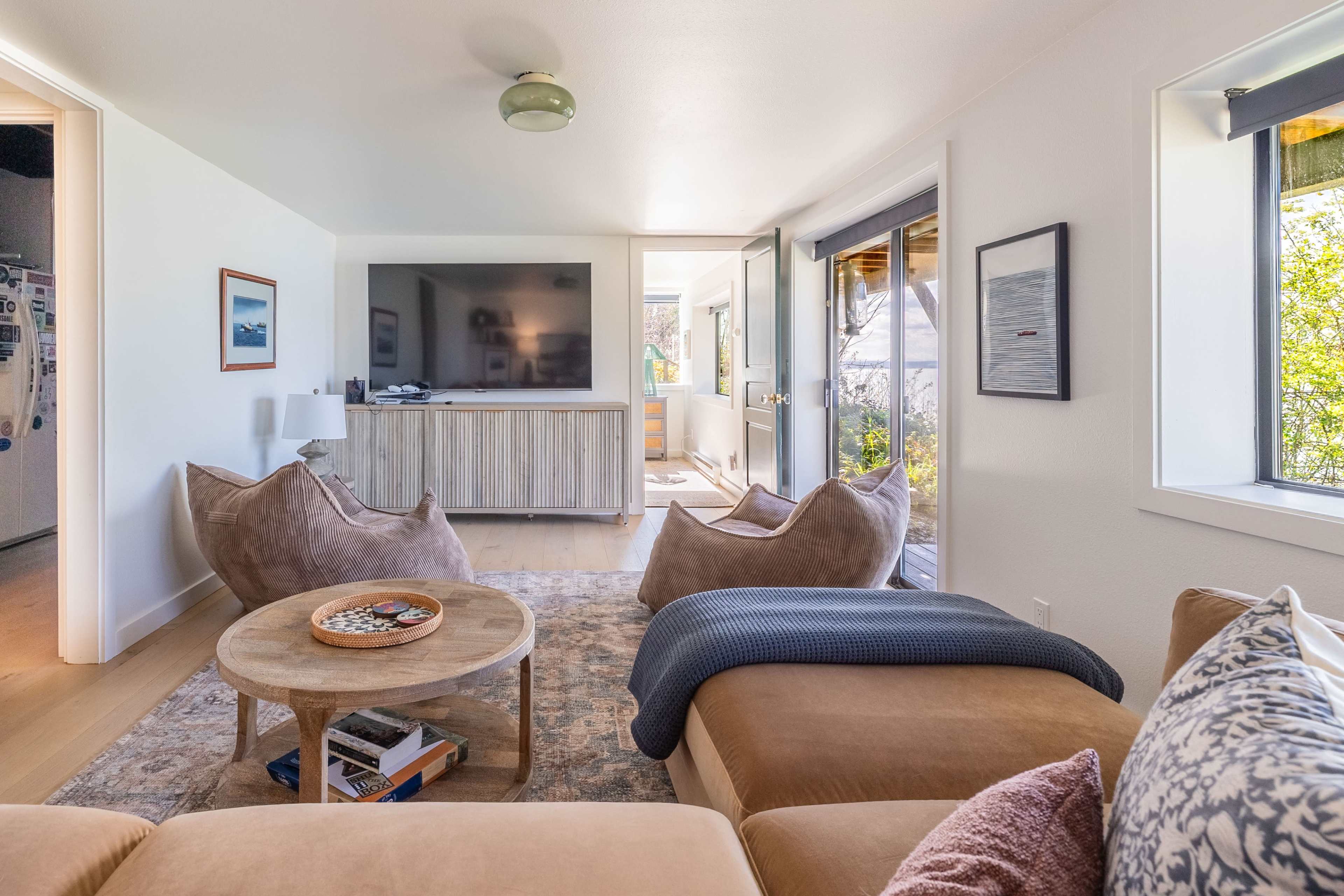 The image shows a modern living room with a large TV, two comfortable armchairs, and a coffee table on a light-colored rug, with sliding doors leading to an outdoor area.