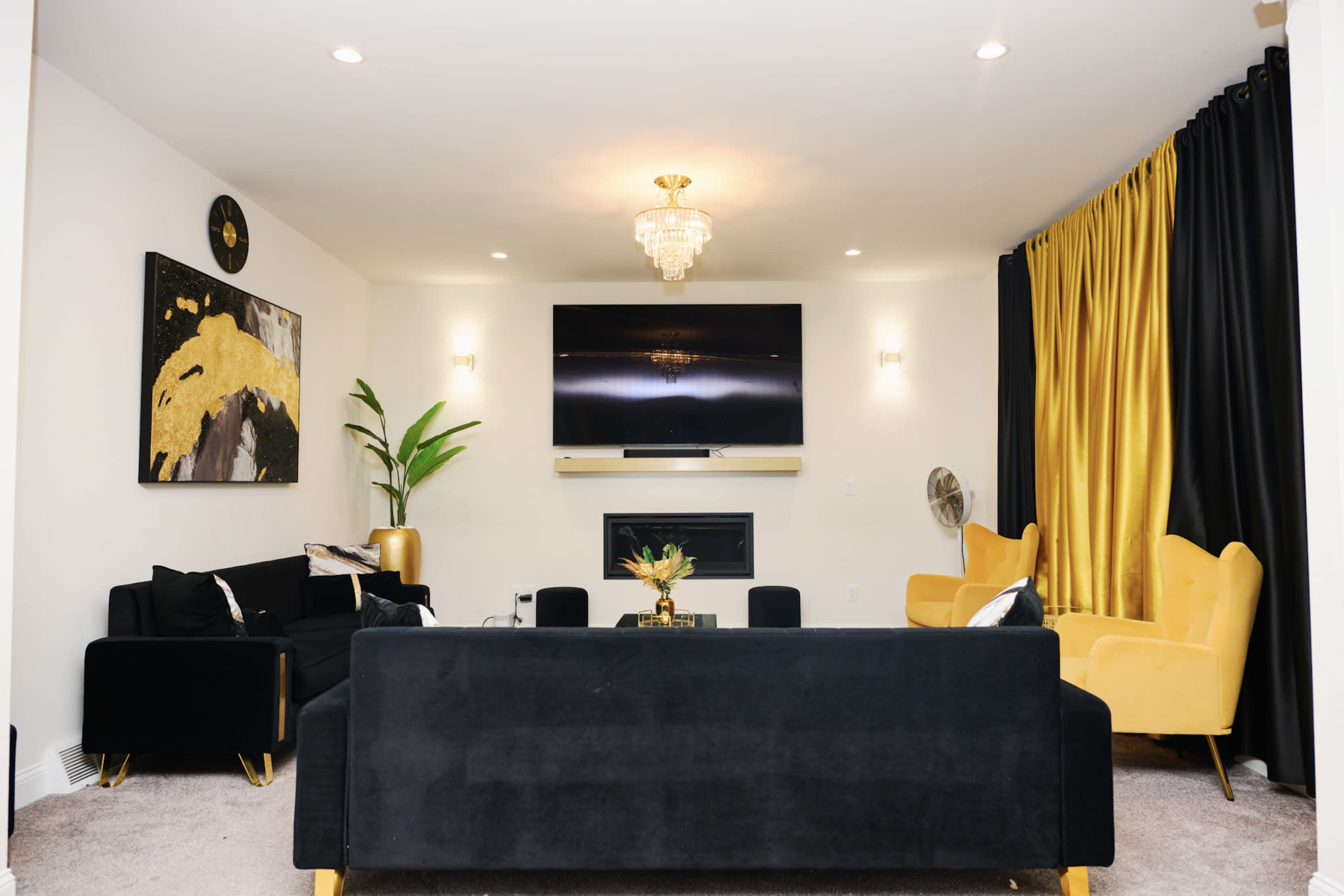 The room features a modern living area with black sofas and chairs, a yellow armchair, a large flat-screen TV mounted above a fireplace, and floor-to-ceiling yellow curtains.