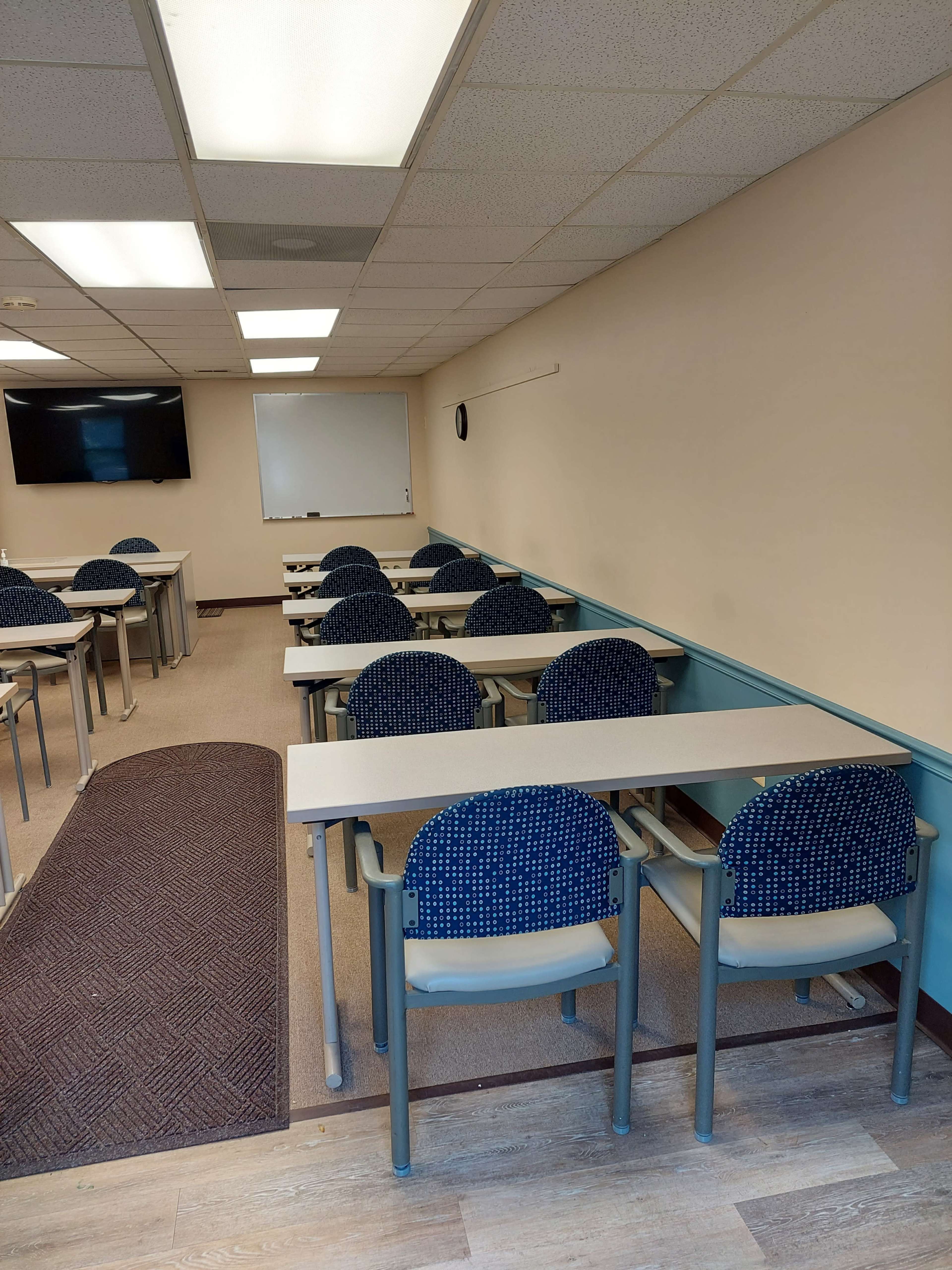 A classroom features rows of desks with blue patterned chairs, a wall-mounted screen, and a whiteboard at the front.