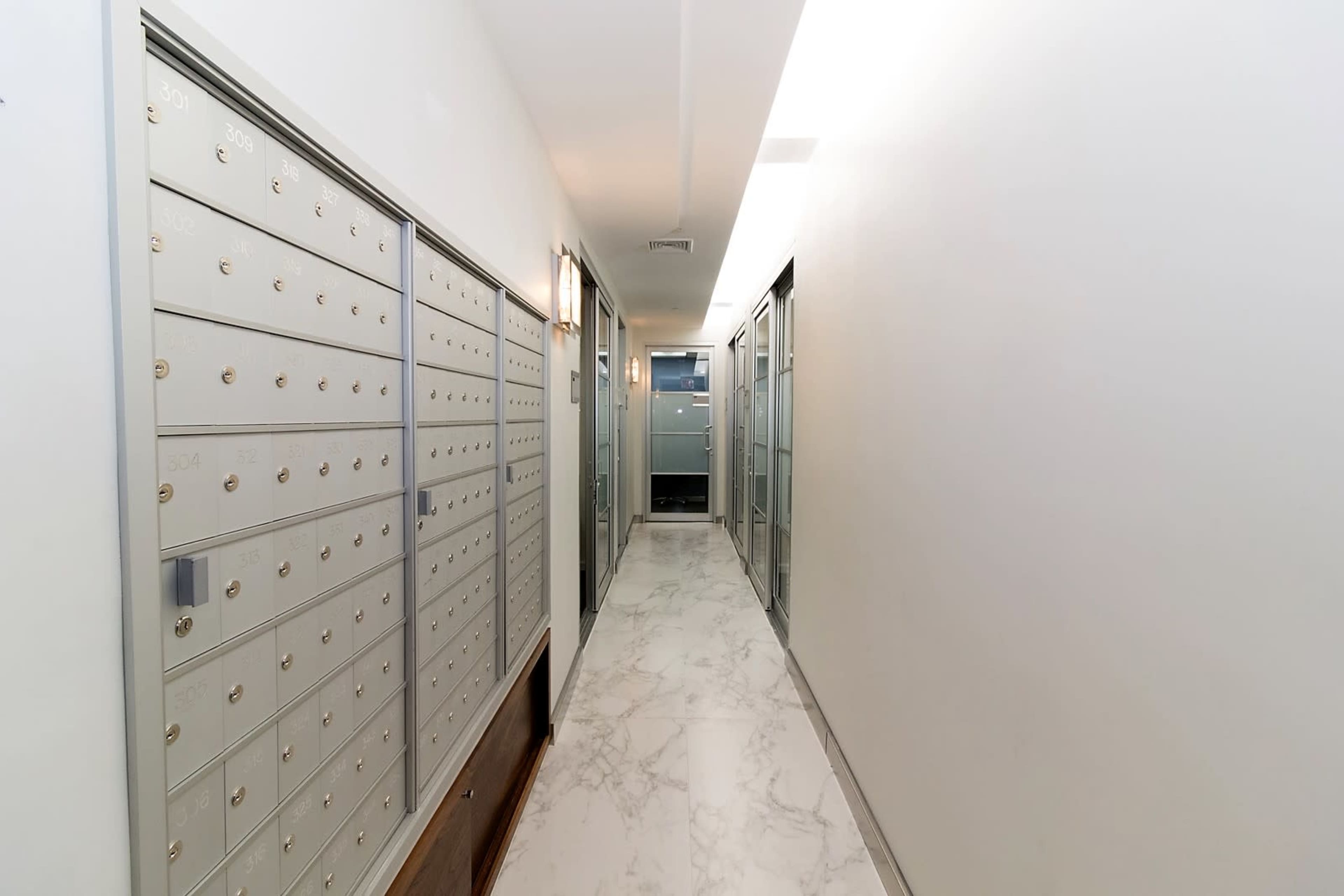 The image shows a hallway lined with mailbox units on one side and glass-fronted doors on the opposite side, featuring a marble floor and modern lighting.