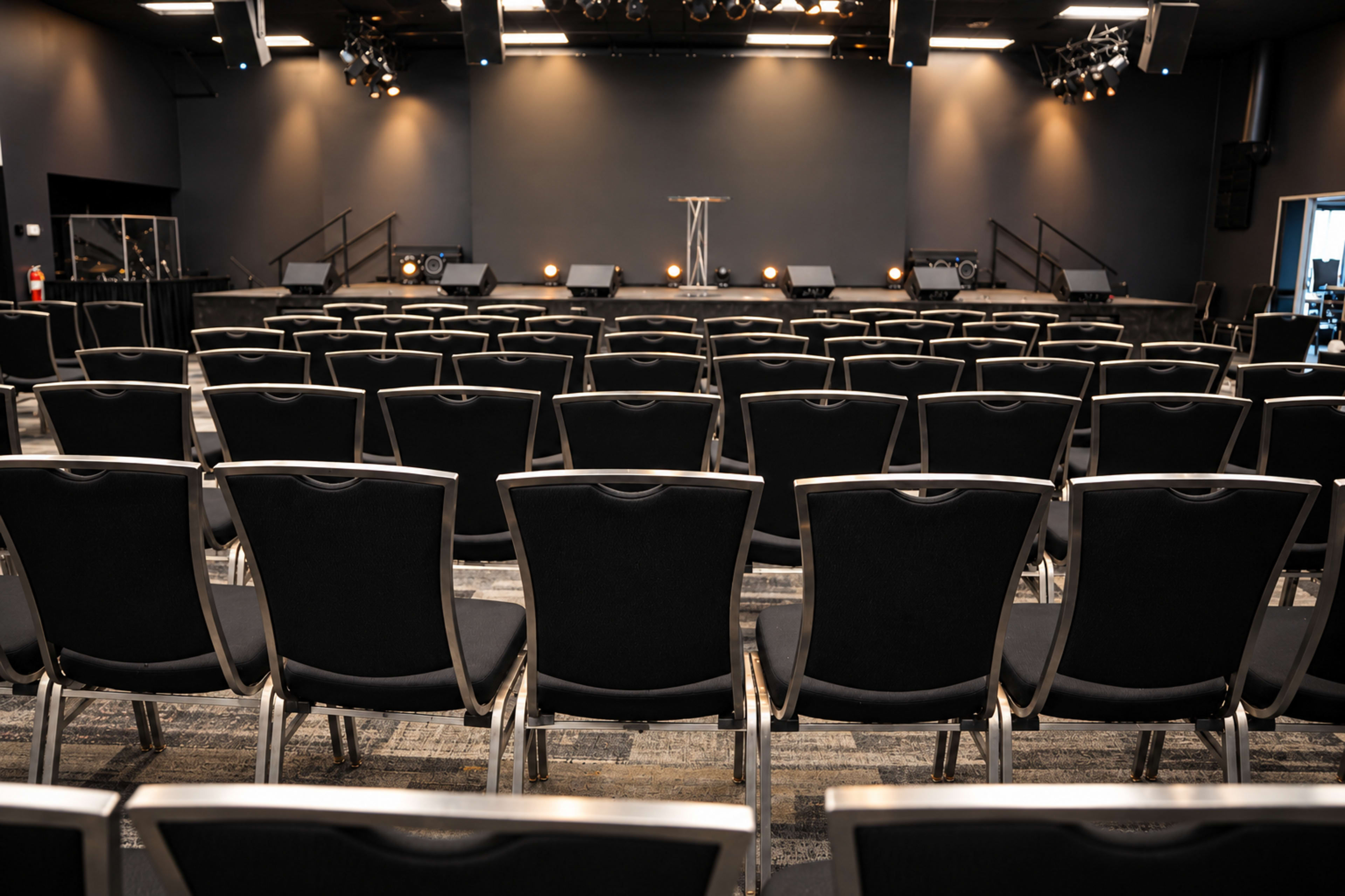 The image shows a spacious auditorium with neatly aligned black chairs facing a stage that is illuminated by spotlights.