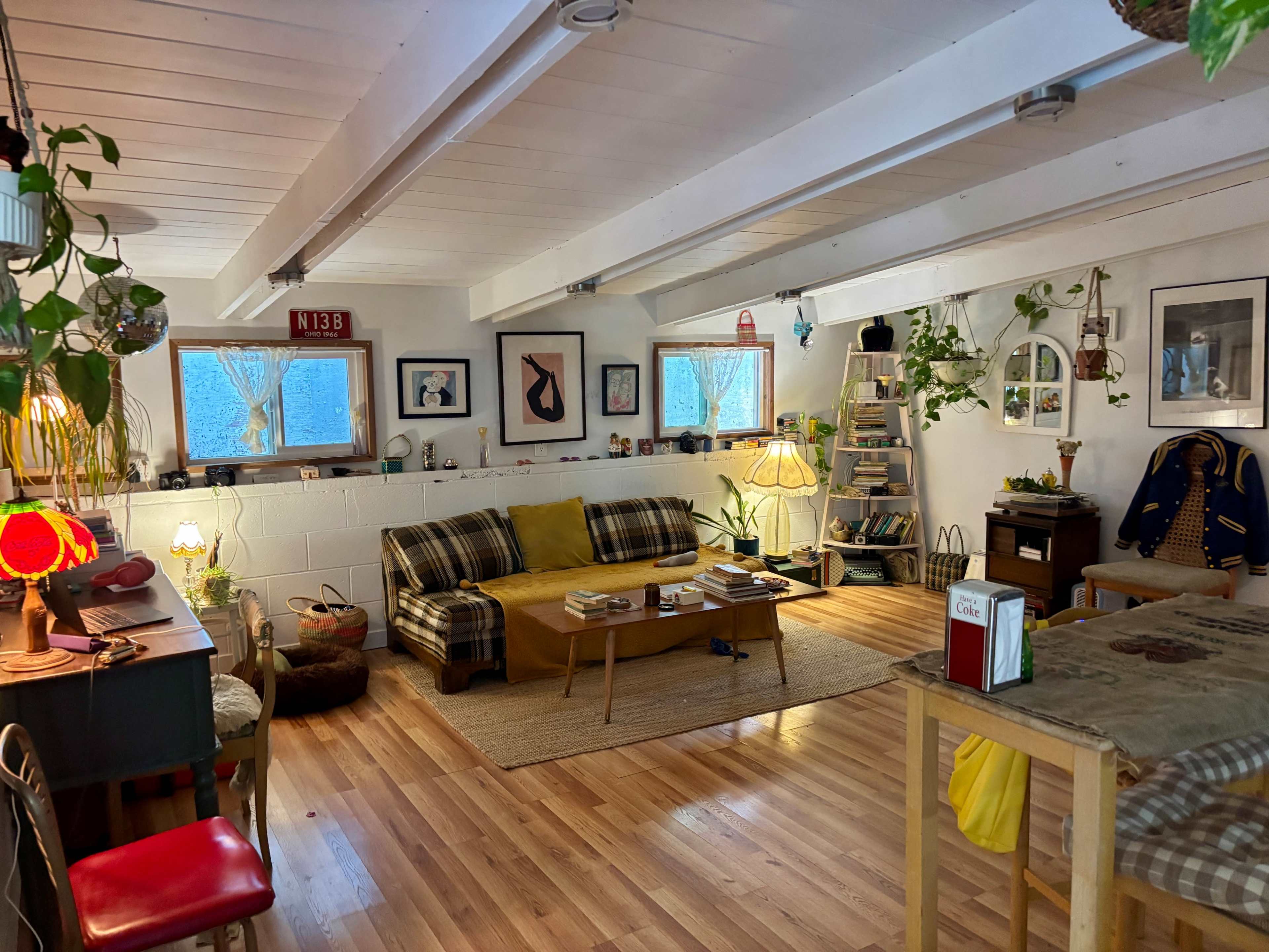 The image shows a spacious, well-lit living area featuring a sofa, wooden coffee table, and various plants, with artwork on the walls and a warm, inviting atmosphere.