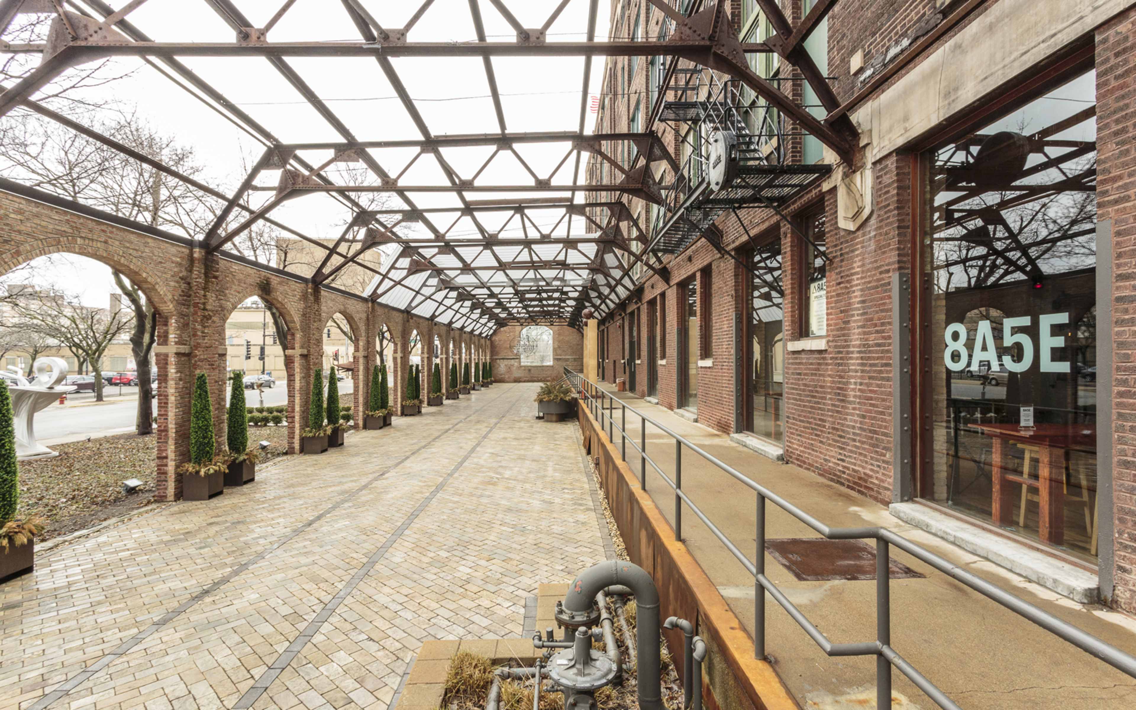 The image shows a brick building with an open walkway covered by a metal framework, lined with small trees and leading to large glass windows.