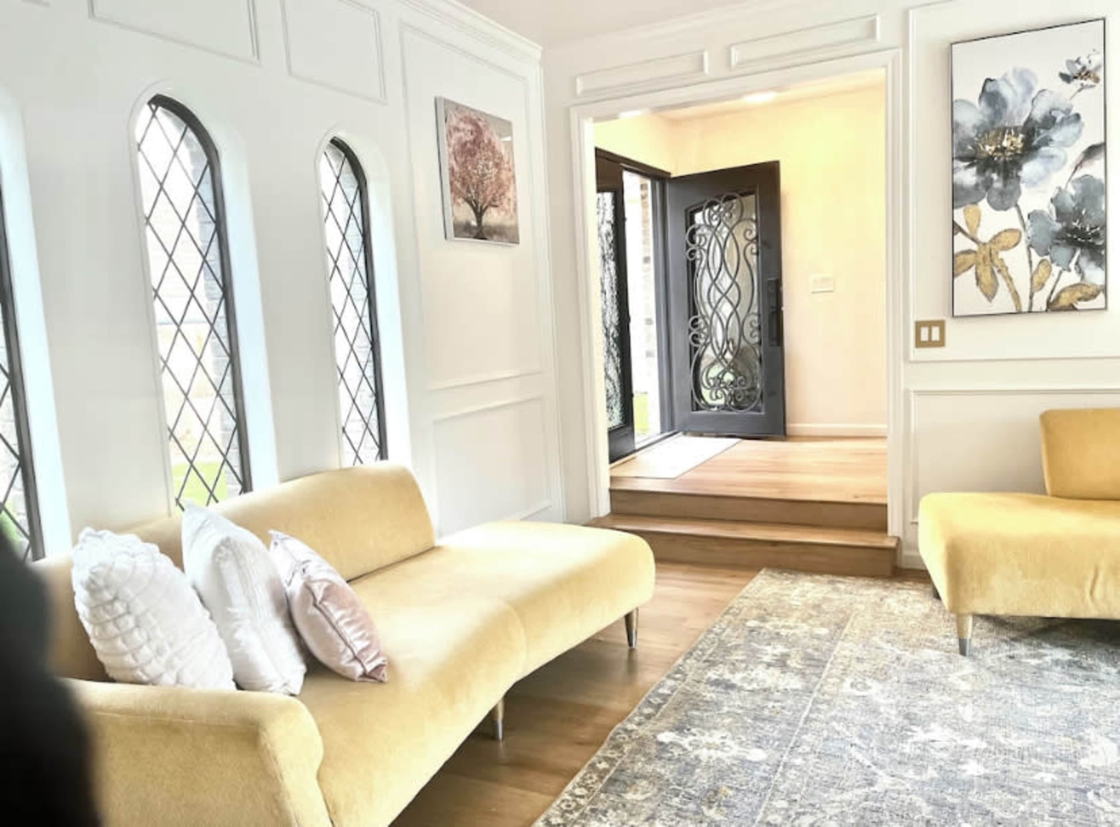 The scene features a light-filled living room with modern furniture, large windows, and a decorative entrance door flanked by steps.