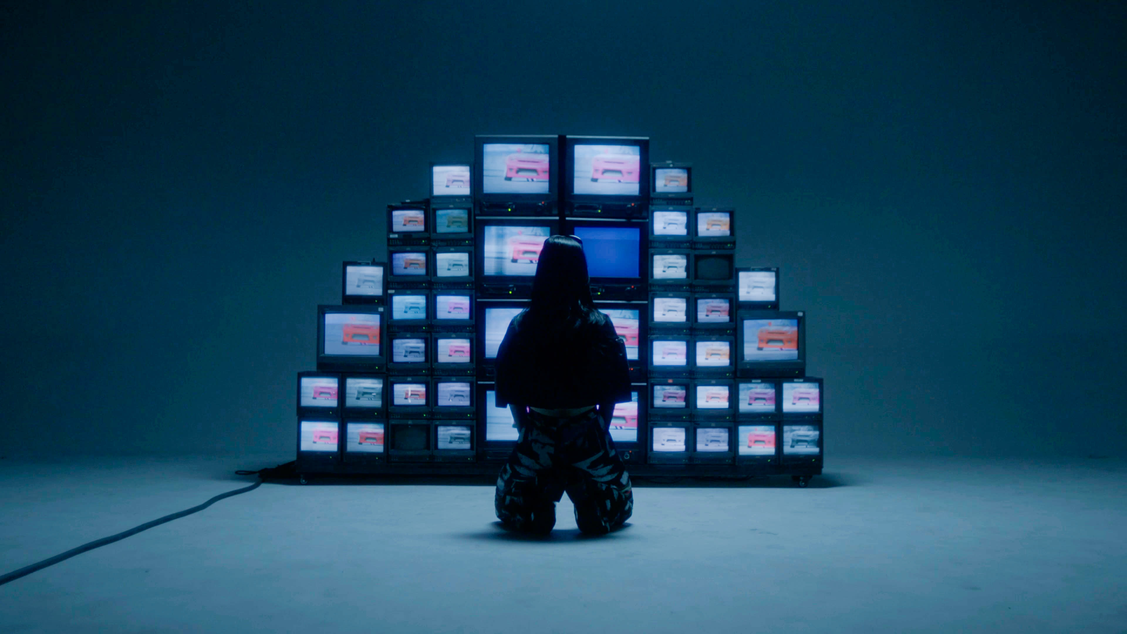 A person kneels in front of a large arrangement of multiple television screens displaying a series of images.