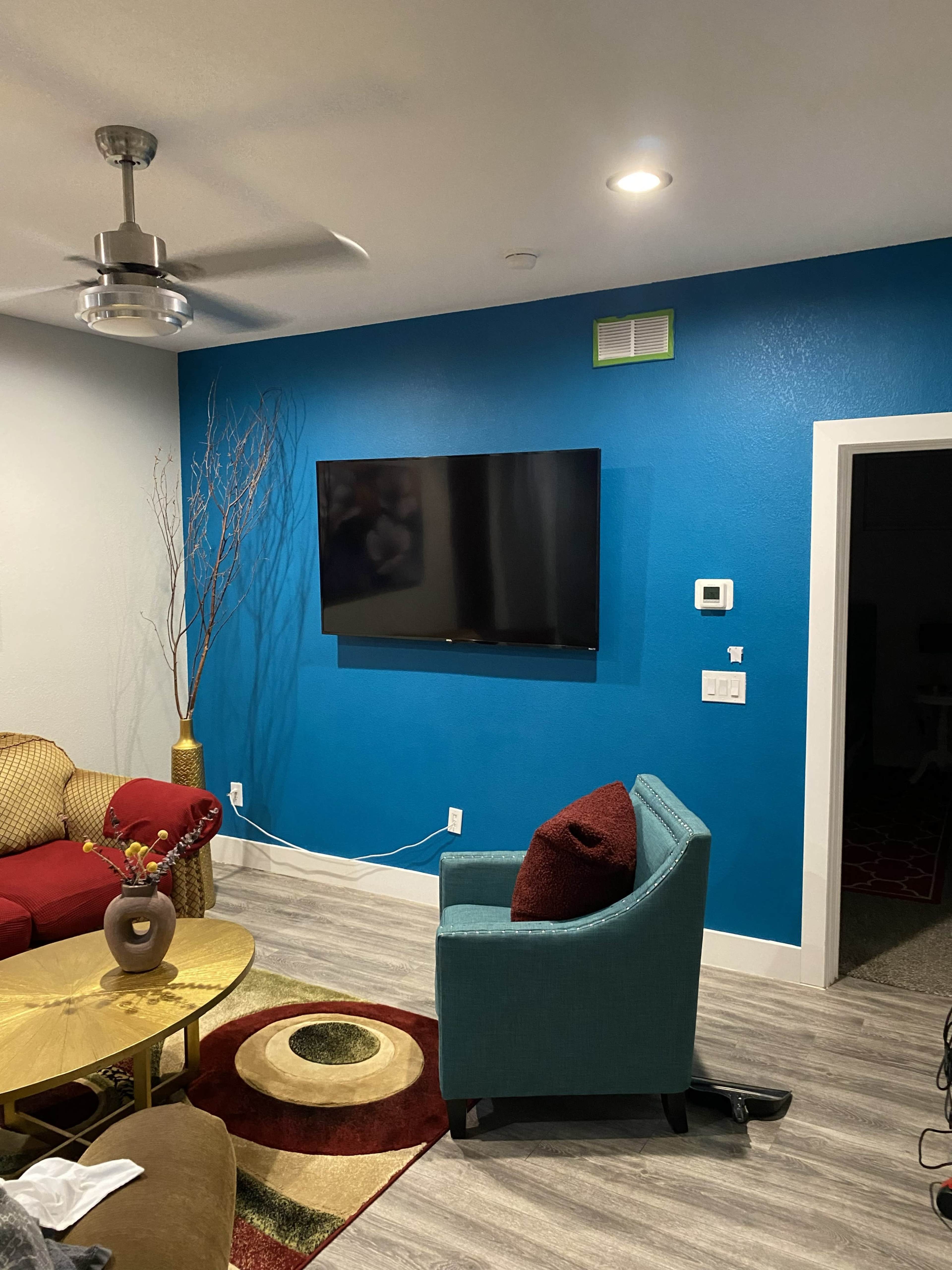 The image shows a living room with a red sofa, teal accent chair, gold coffee table, and a wall-mounted TV against a blue wall.