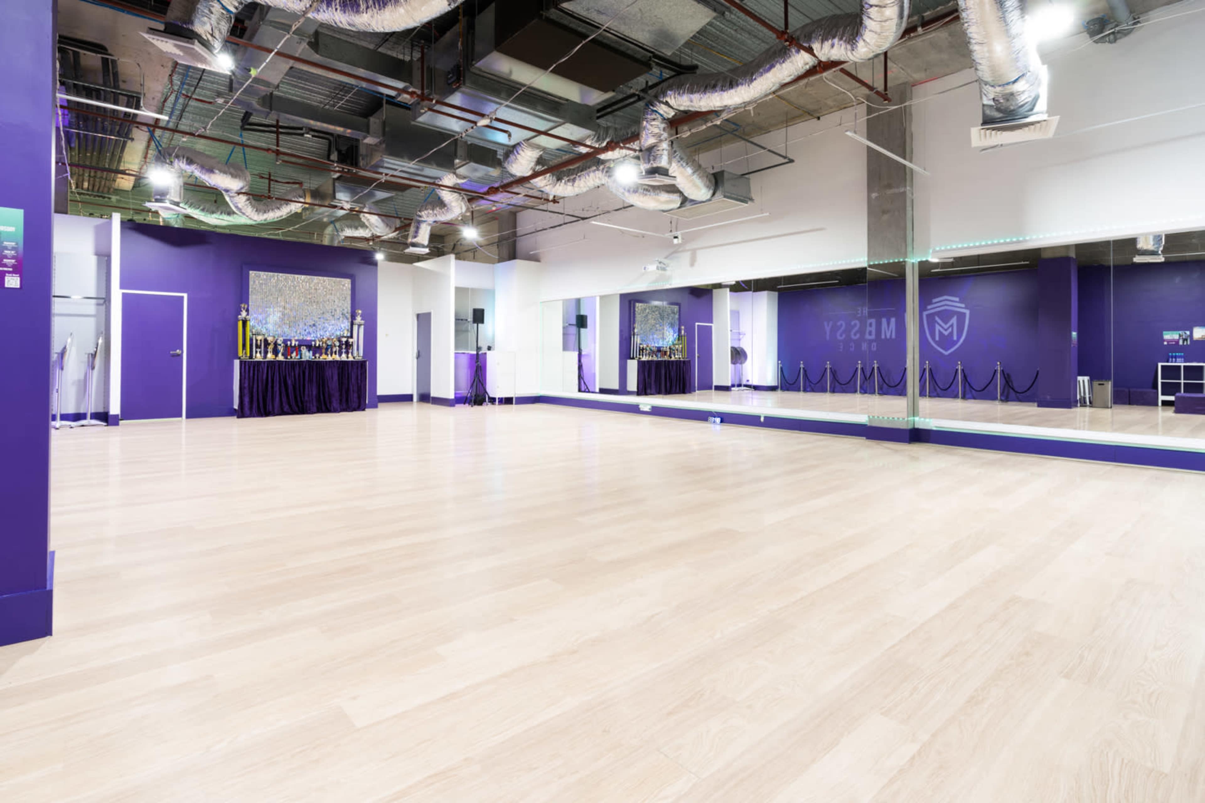 The image shows a spacious, empty dance studio with light wooden flooring, purple walls, and large mirrors.