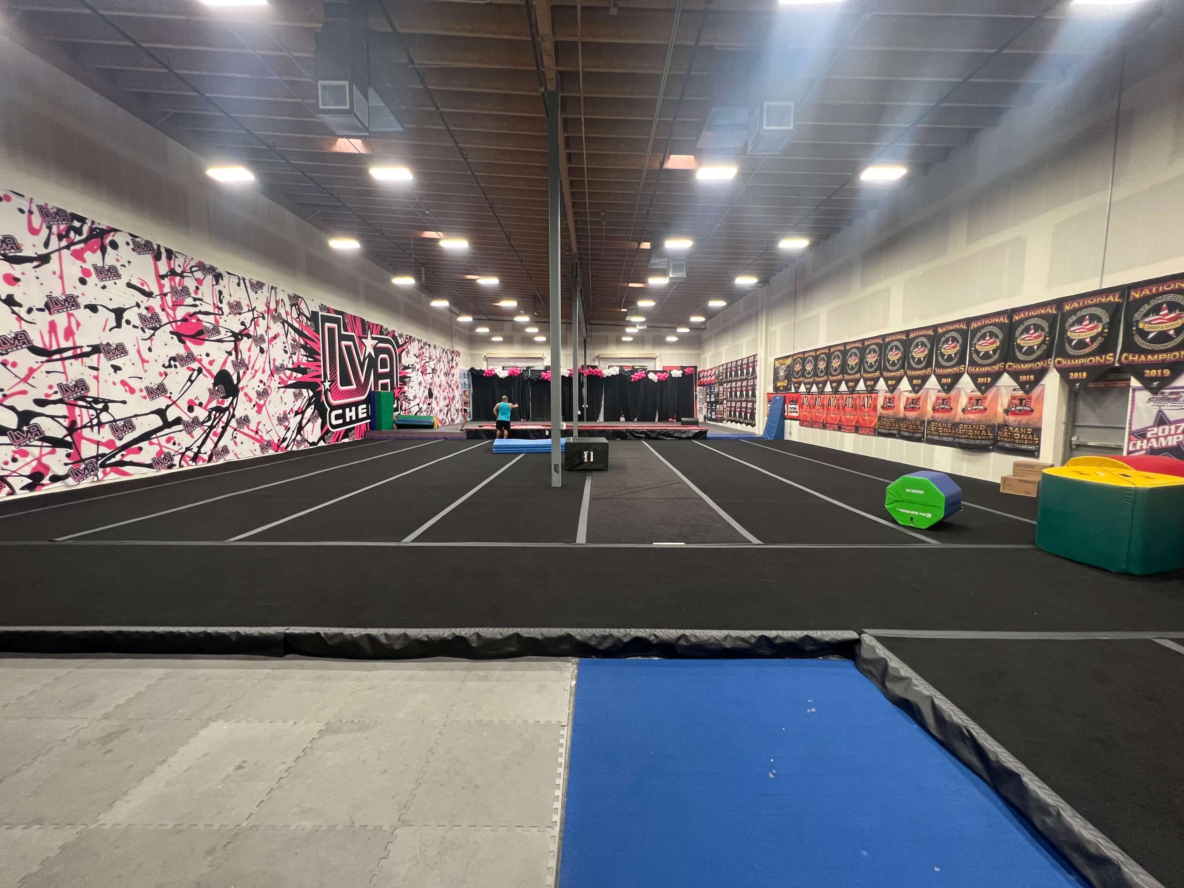 The image shows an indoor gymnastics facility with black flooring, framed by a colorful wall mural and lined with various training equipment along the sides.