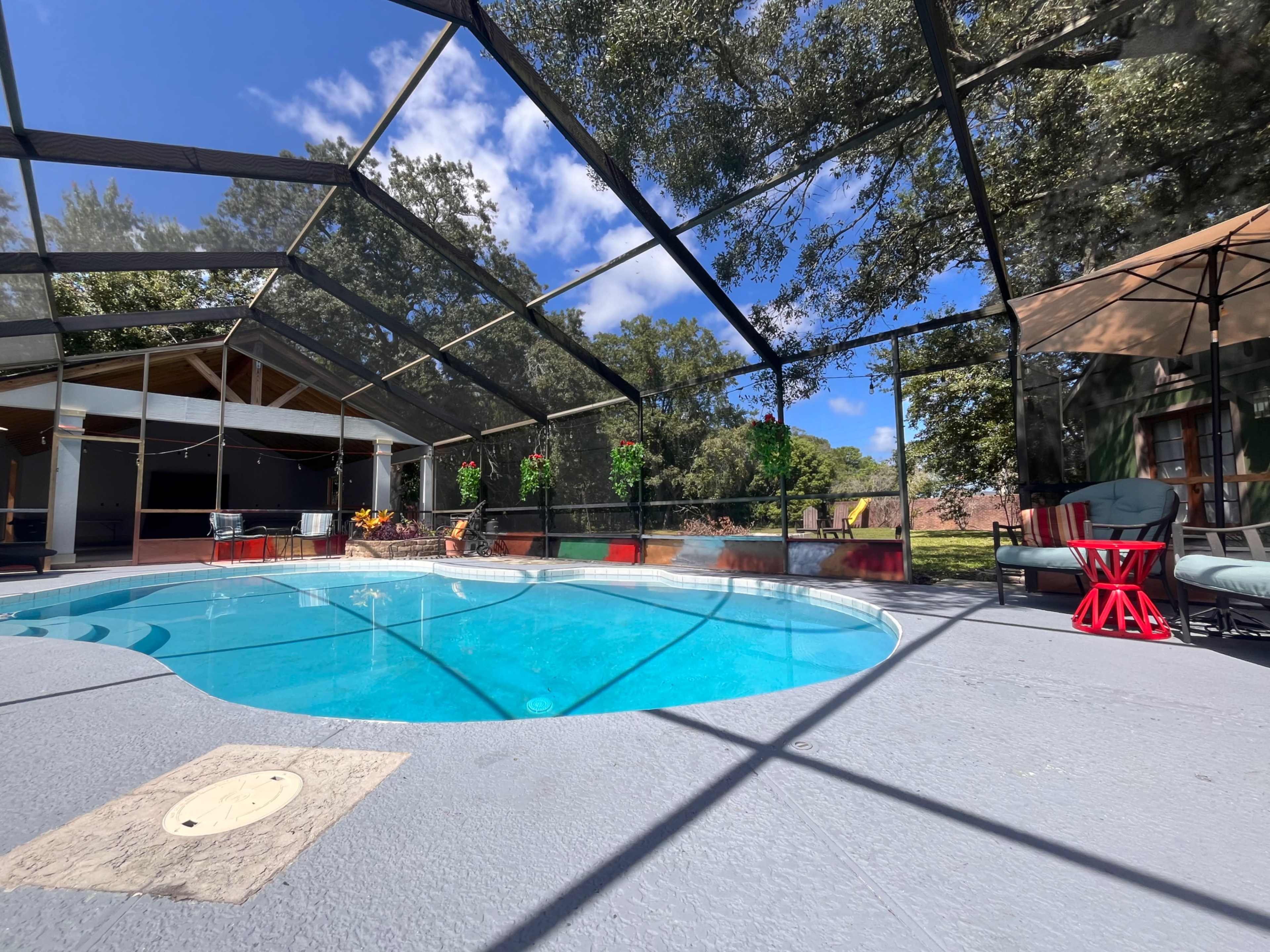 The image shows a blue swimming pool enclosed by a screened-in patio, surrounded by green trees and a clear blue sky.