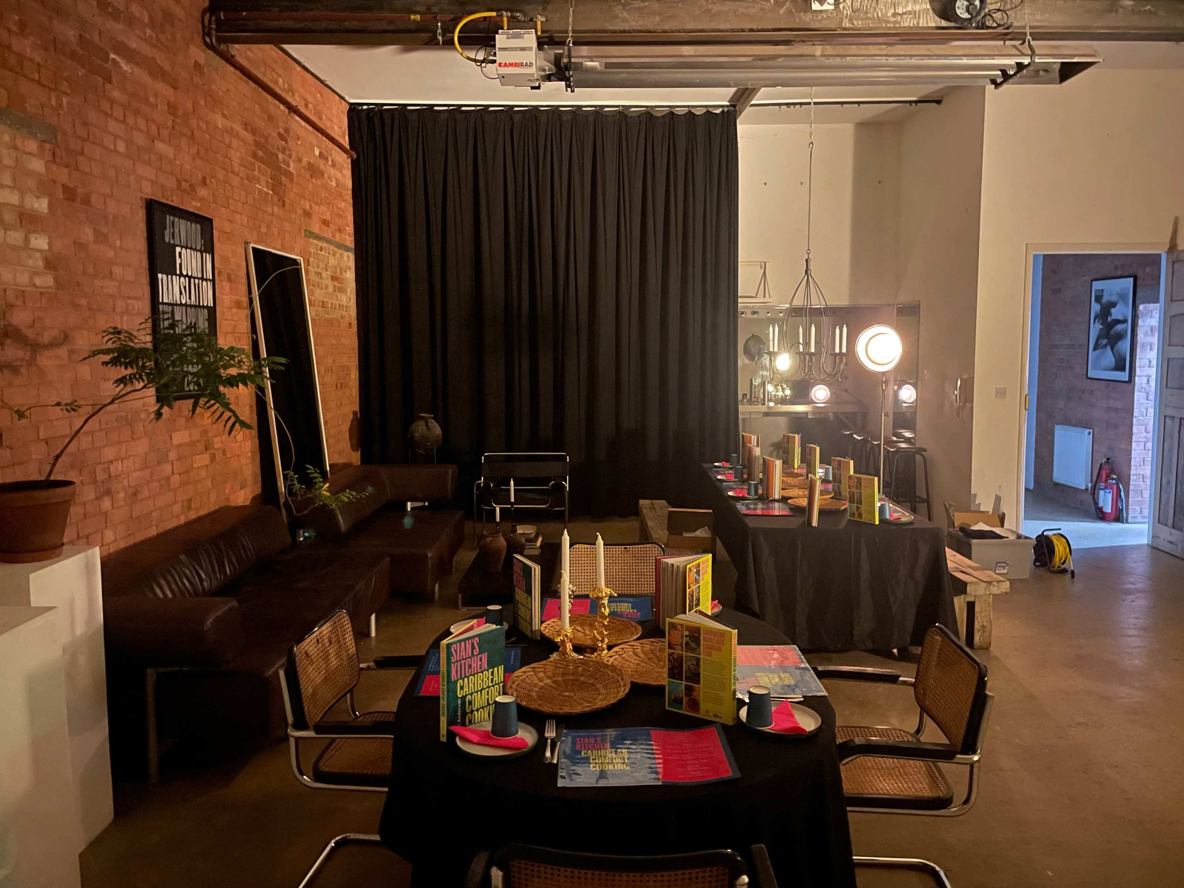 The image shows a stylish event space with a brick wall, tables set with colorful materials, and a dark curtained area in the background.