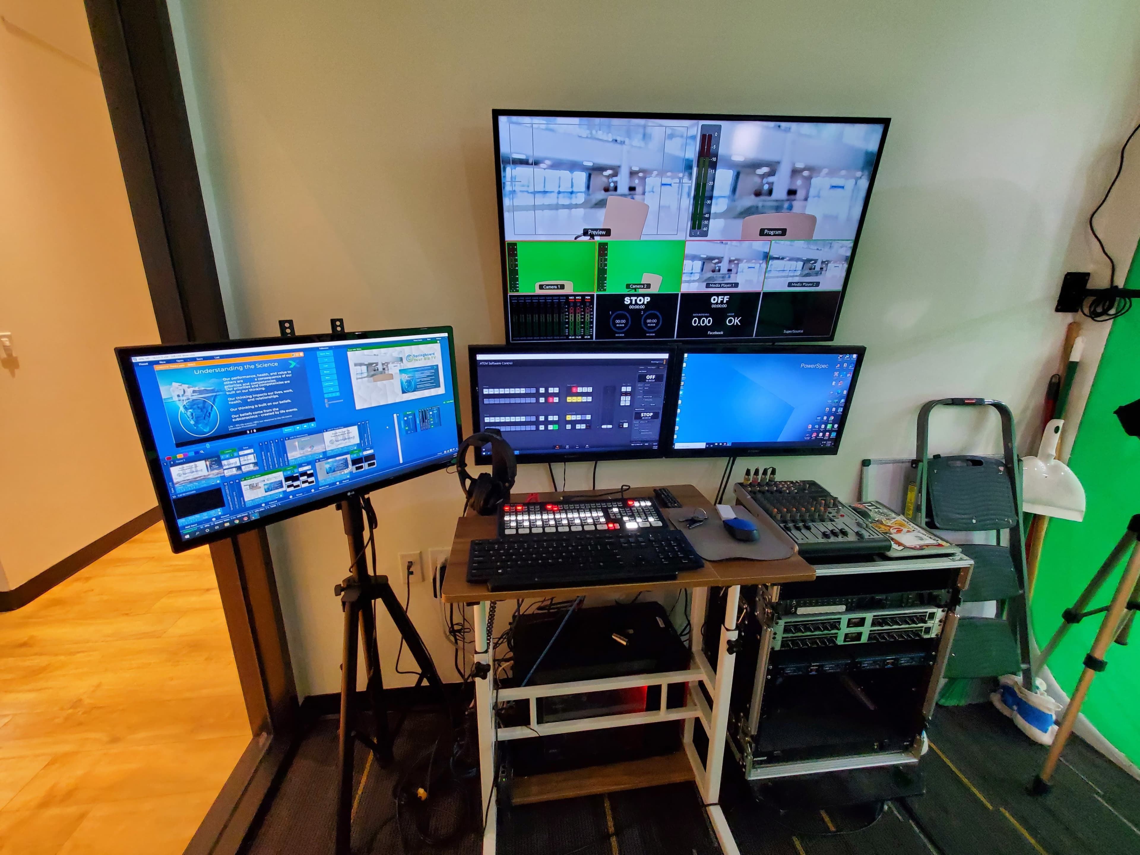 The image shows a multimedia control station featuring multiple monitors, a keyboard, audio mixer, and various equipment arranged on a mobile cart against a wall.
