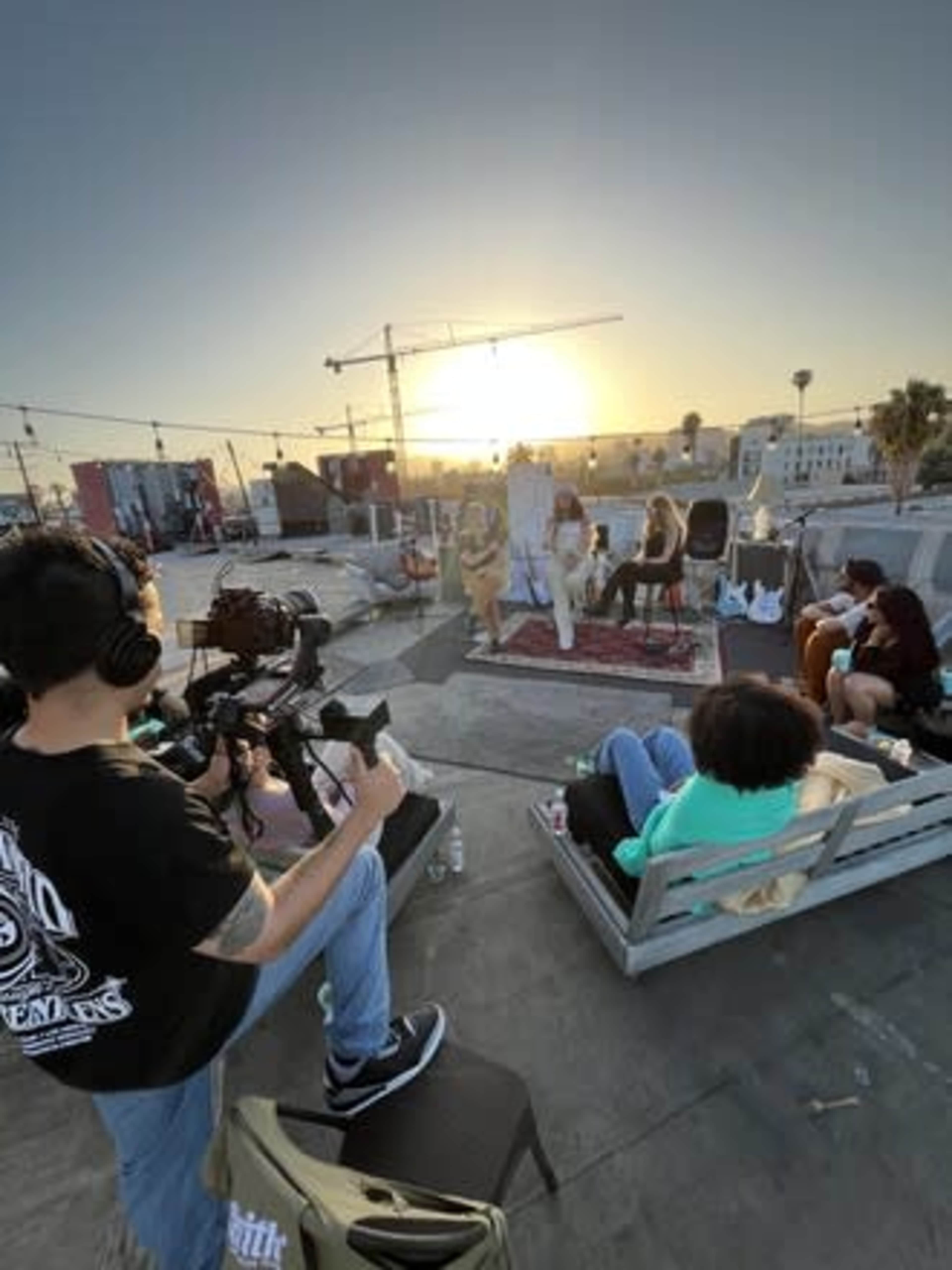 A film crew records a performance on a rooftop as the sun sets in the background, with several people seated in casual seating arrangements.