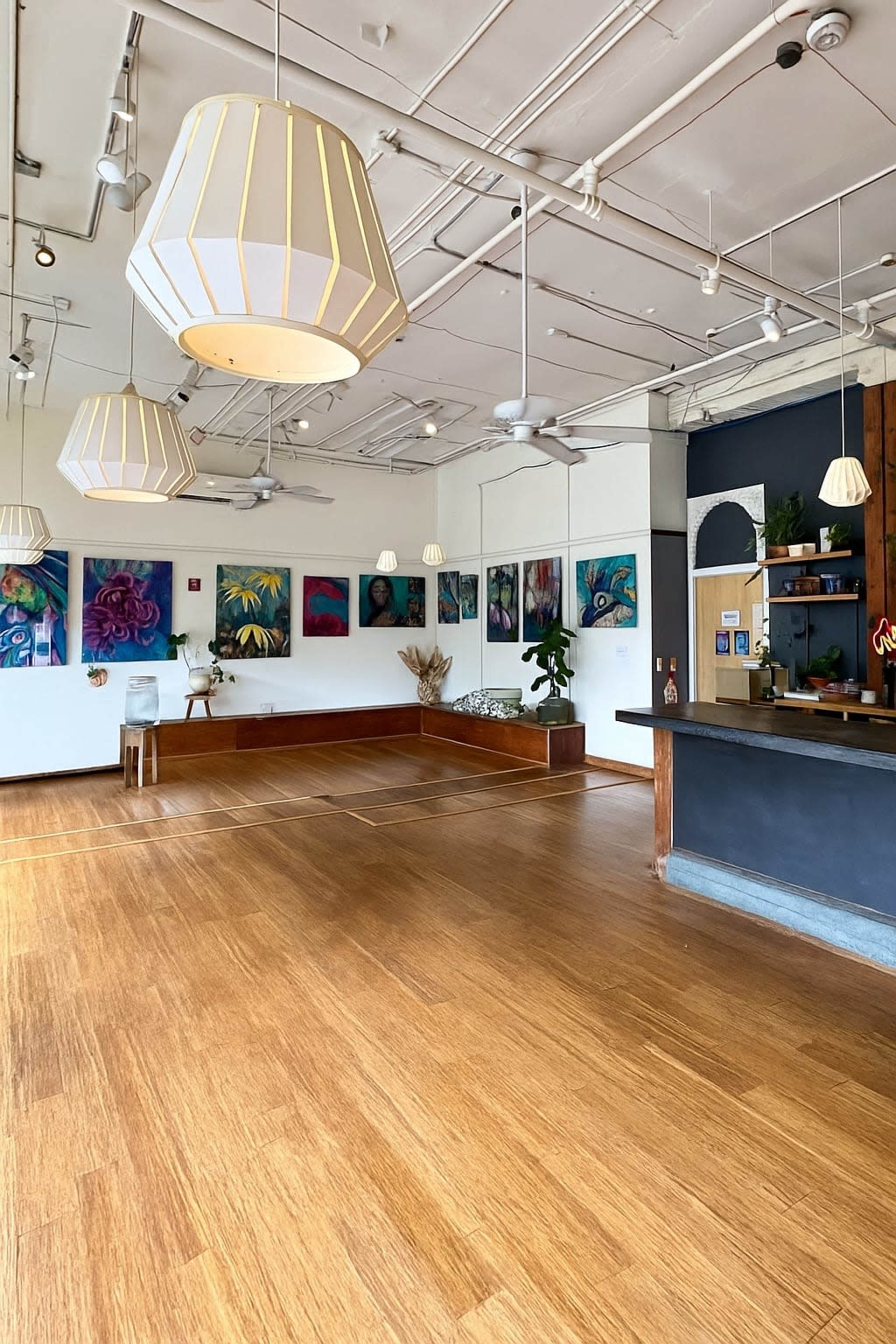 The image shows a spacious interior with wooden flooring, pendant lights, colorful artwork on the walls, and a seating area alongside a counter.