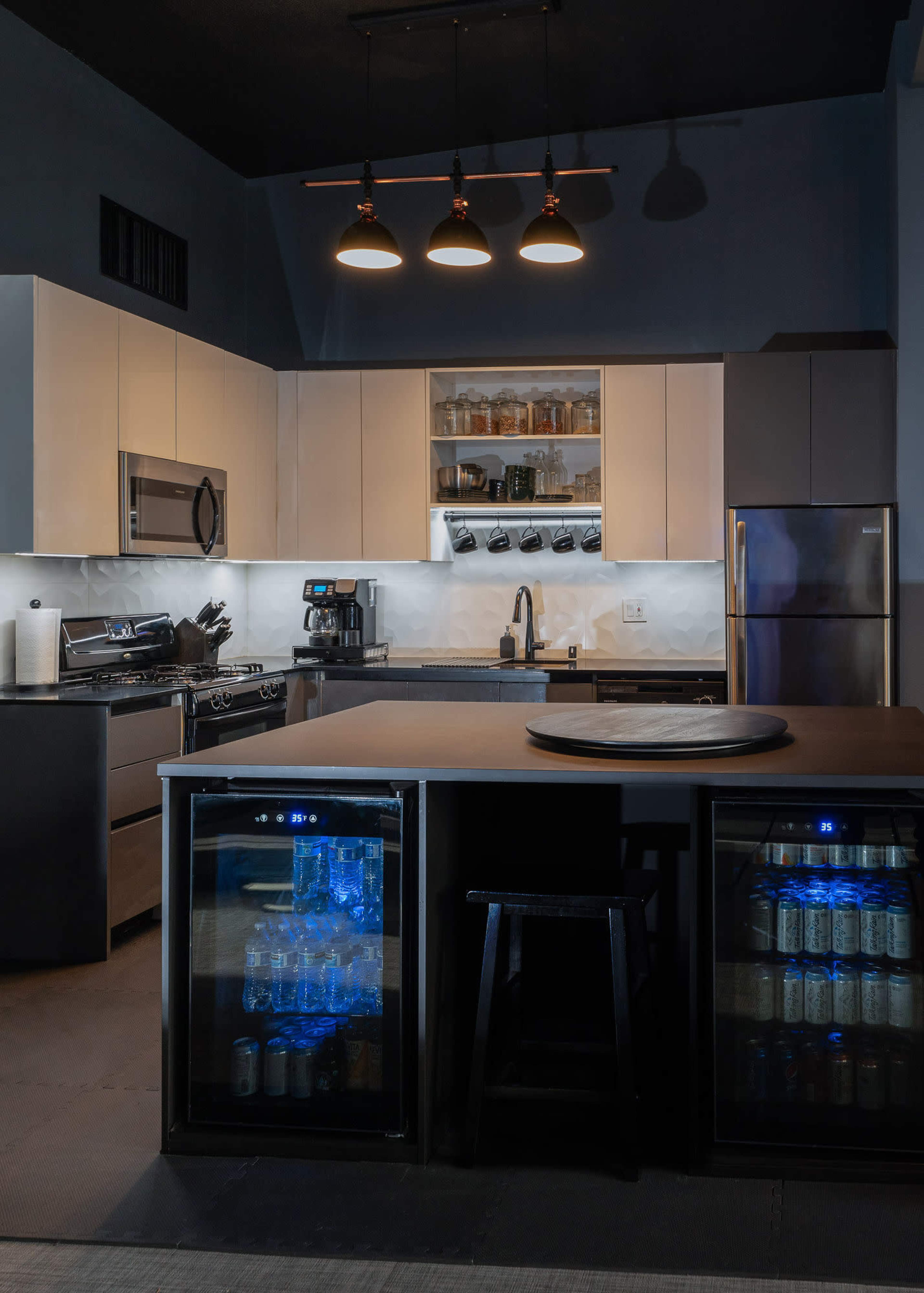 The kitchen features modern appliances and a central island with glass-fronted refrigerators showcasing beverages.