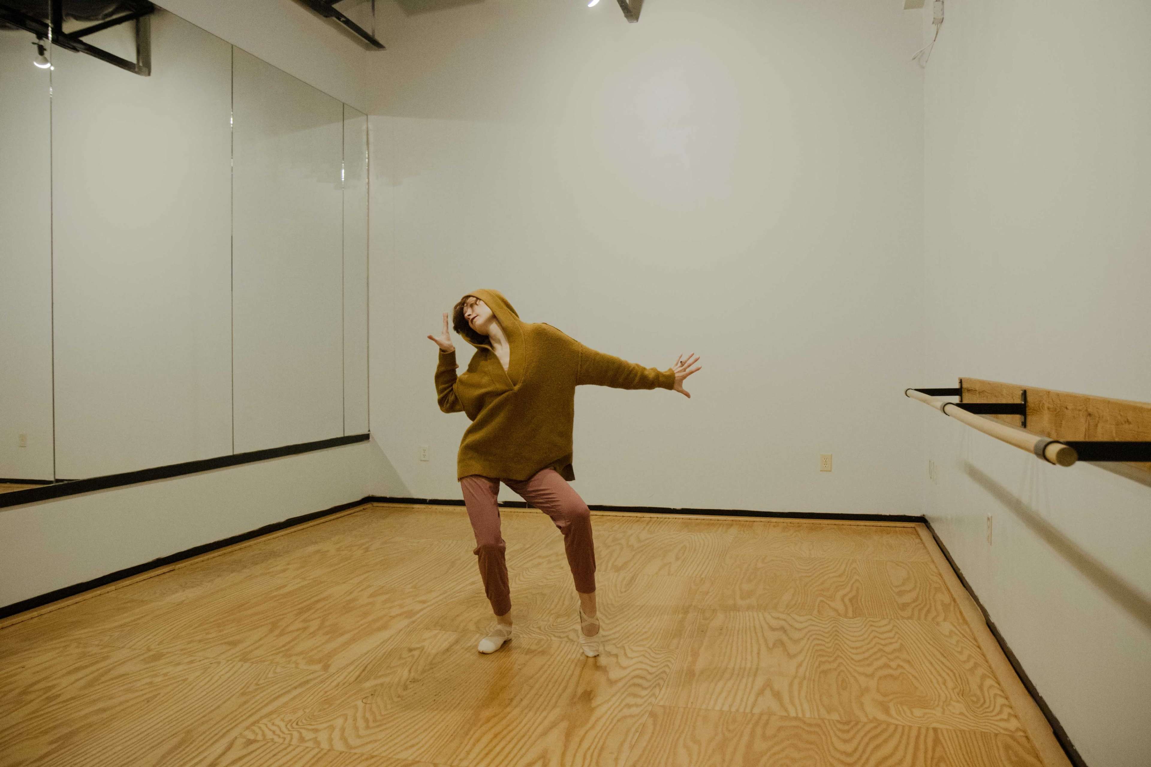 A person in a brown hoodie and pink pants dances in a mirror-lined studio with wooden flooring.