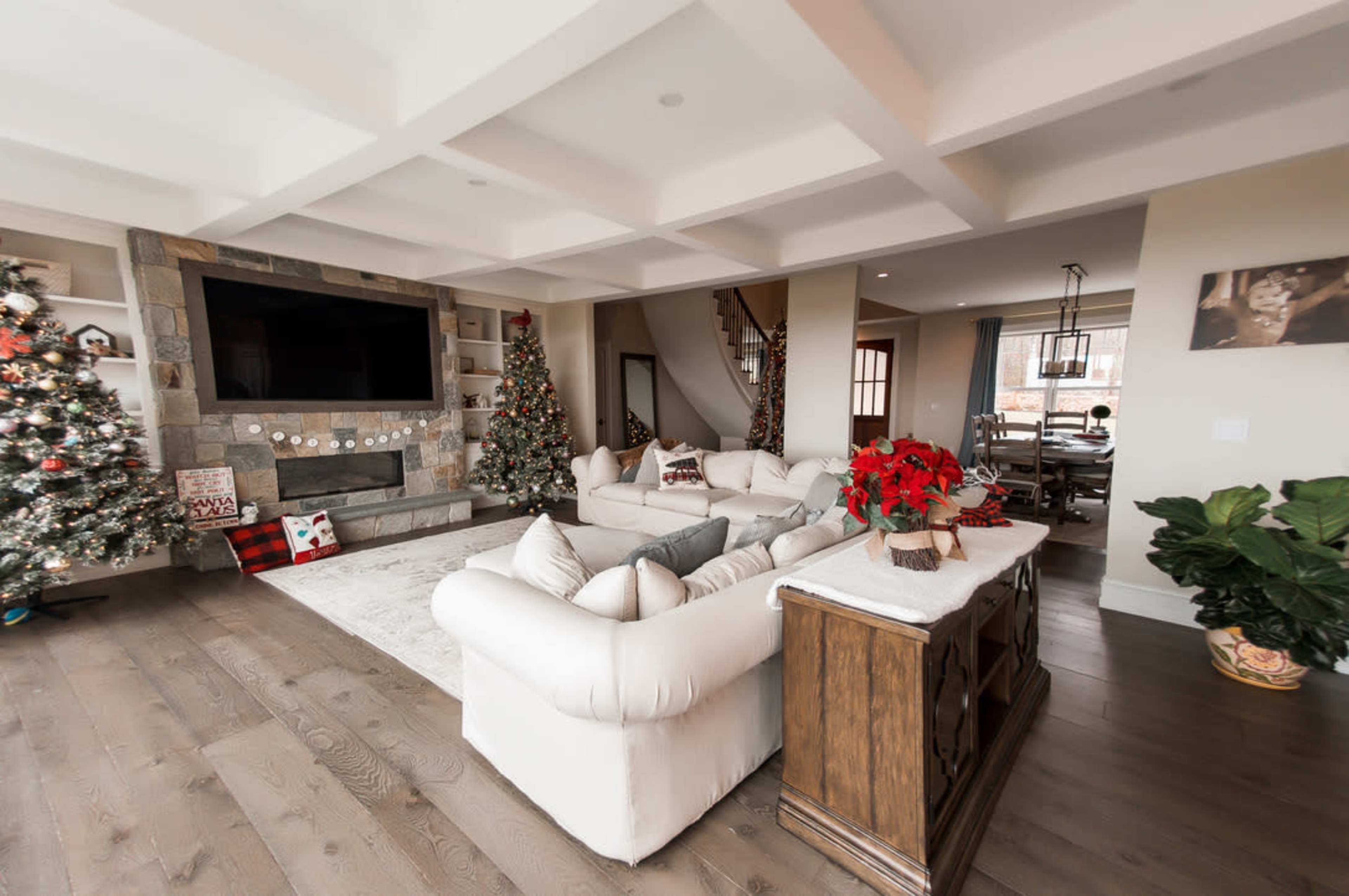 A spacious living room adorned with two Christmas trees, a large television mounted on the wall, and a decorative wooden cabinet.