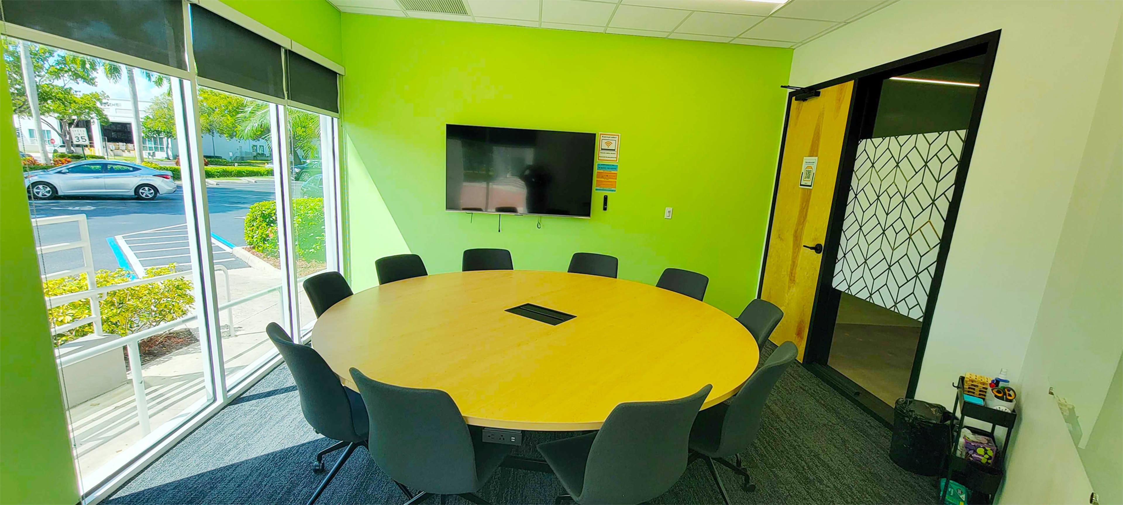 A round conference table surrounded by chairs is set in a brightly lit meeting room with a large window and a wall-mounted television.