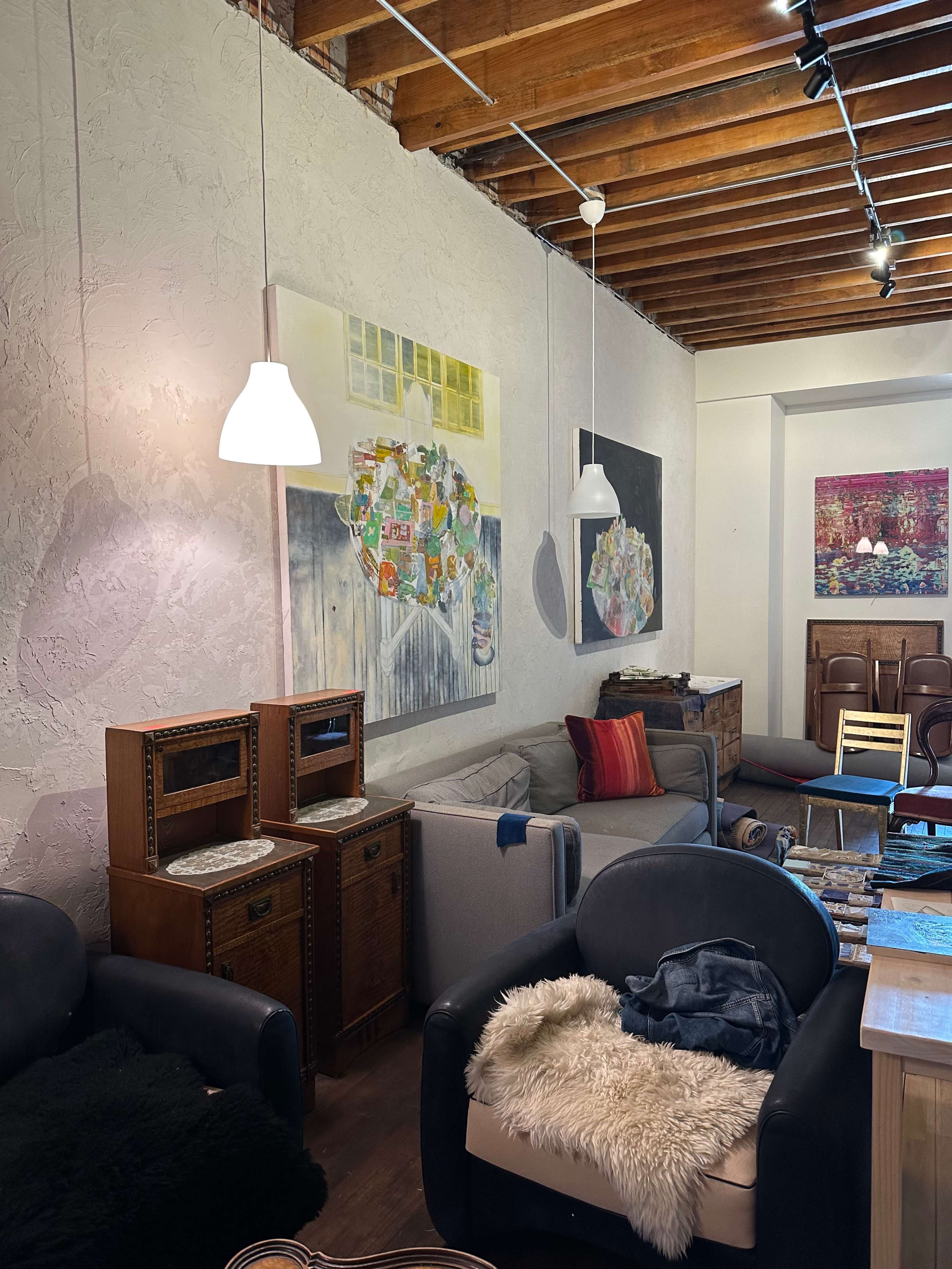 The scene shows a cozy interior with two upholstered armchairs, a gray sofa, wooden furniture, and colorful artworks displayed on the walls.