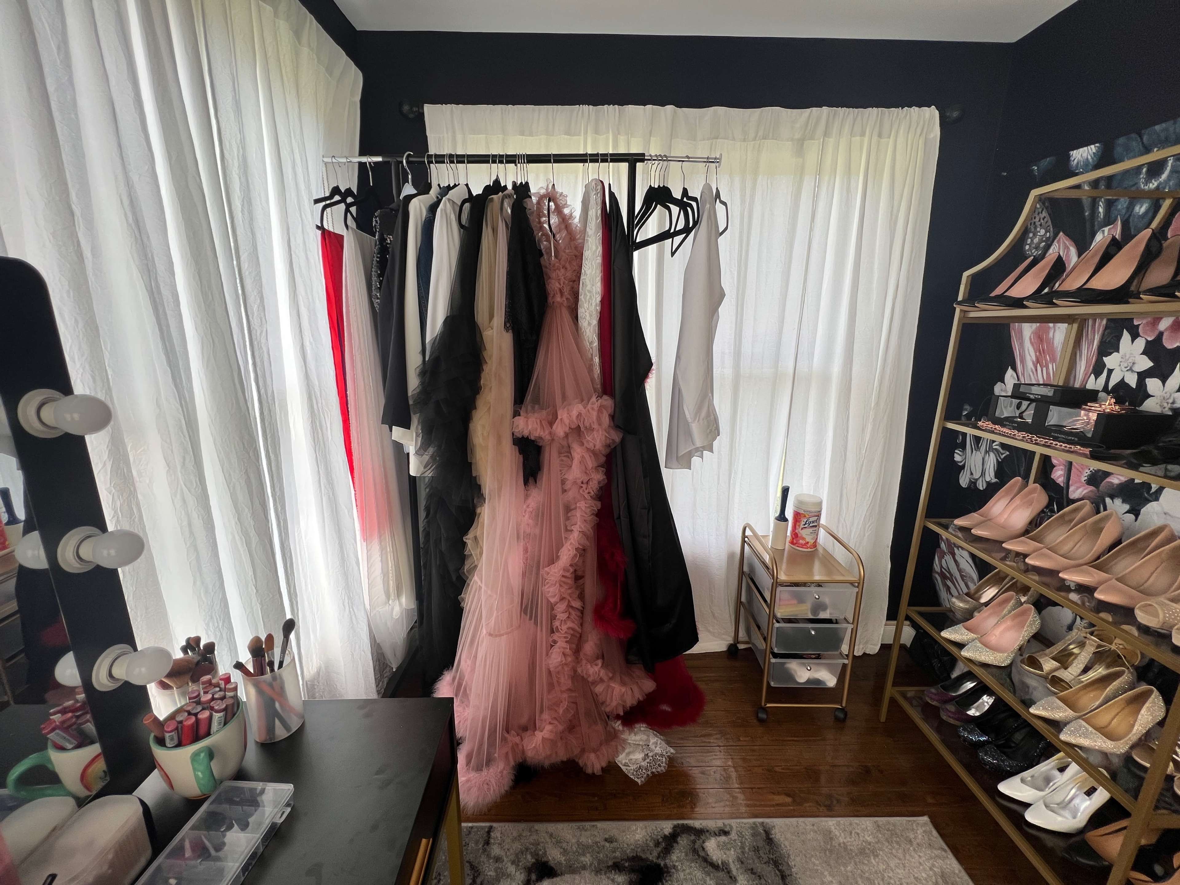 A clothing rack with various dresses and tops stands beside a shelf filled with shoes in a well-lit room featuring large windows.