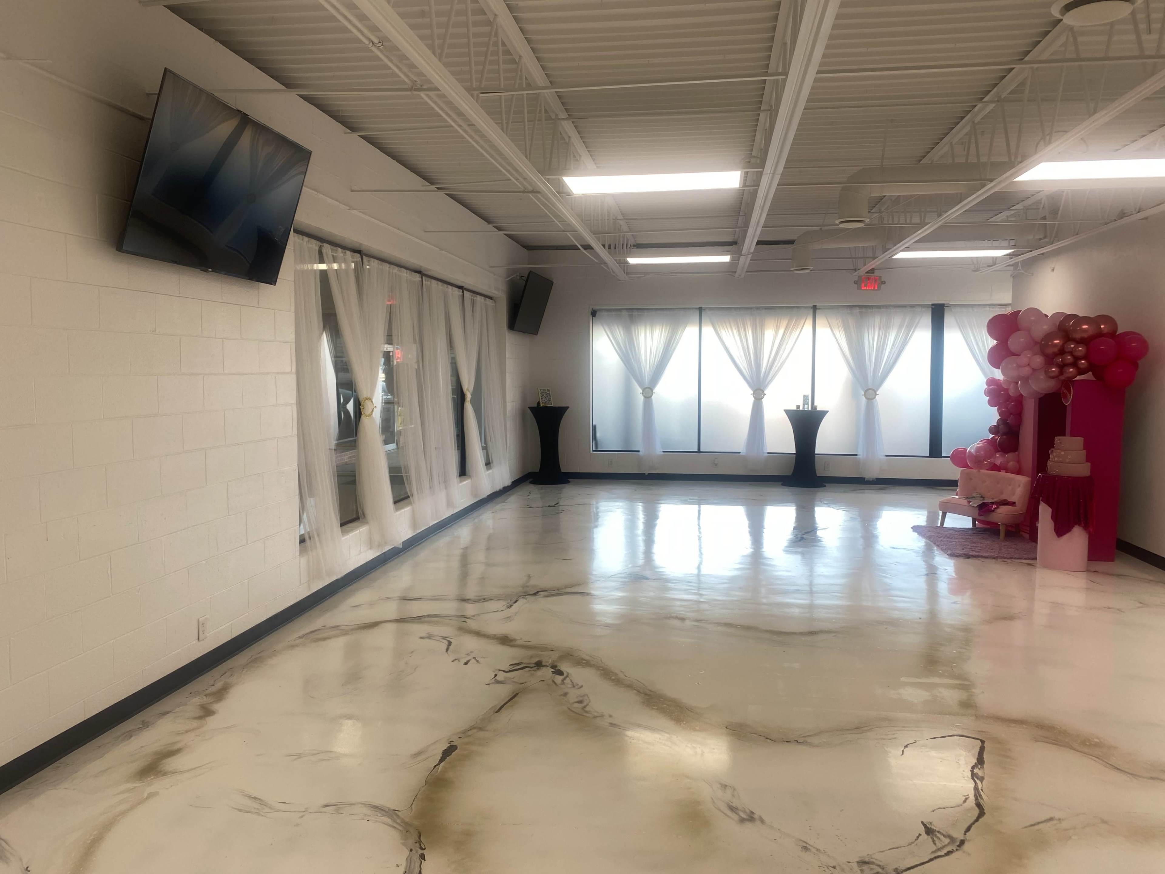 A spacious room with a polished floor, large windows covered by sheer curtains, and decorative elements including pink balloons and a display area at one end.