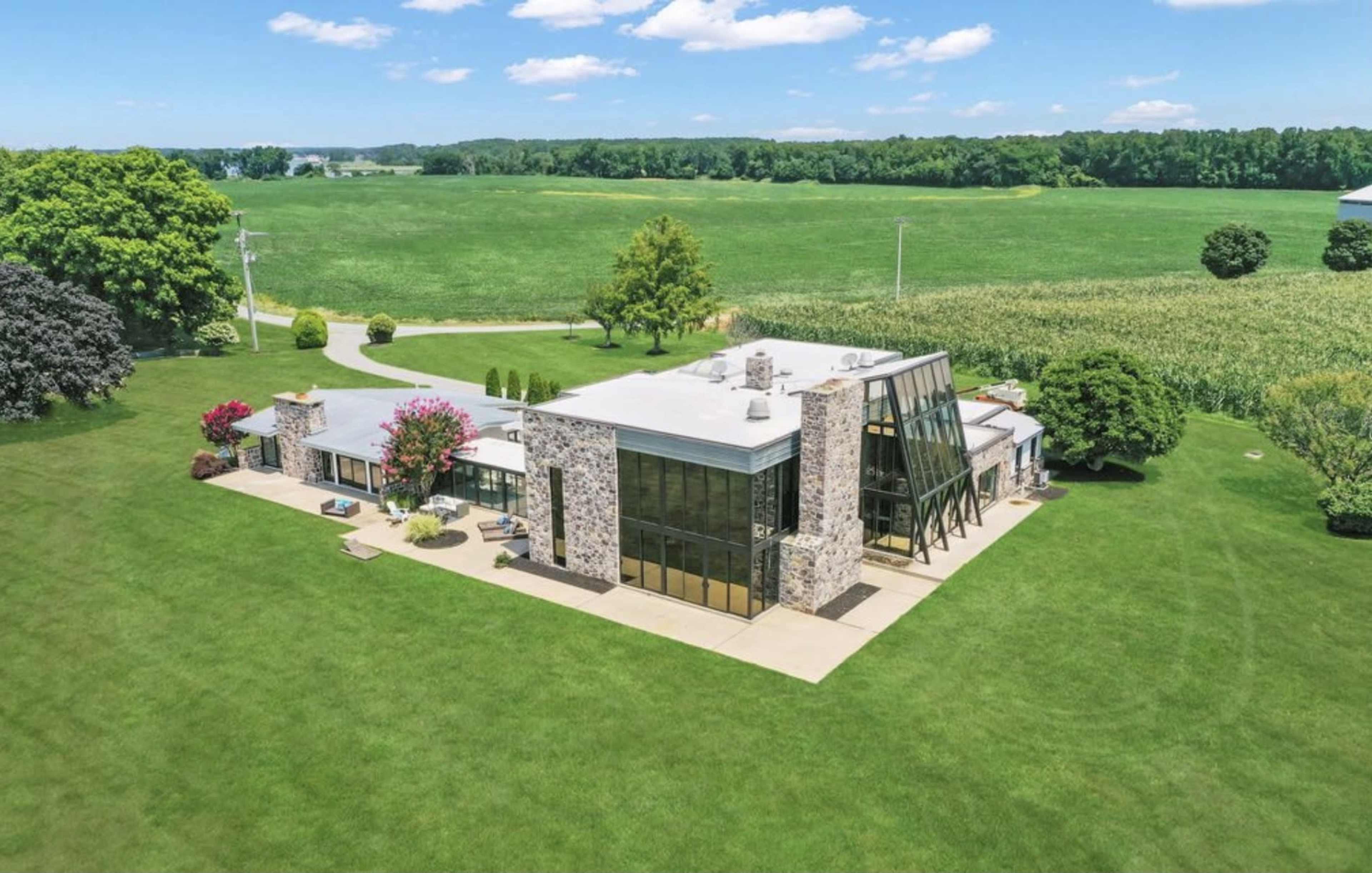 A modern stone and glass home sits in a green landscape with fields and trees nearby.