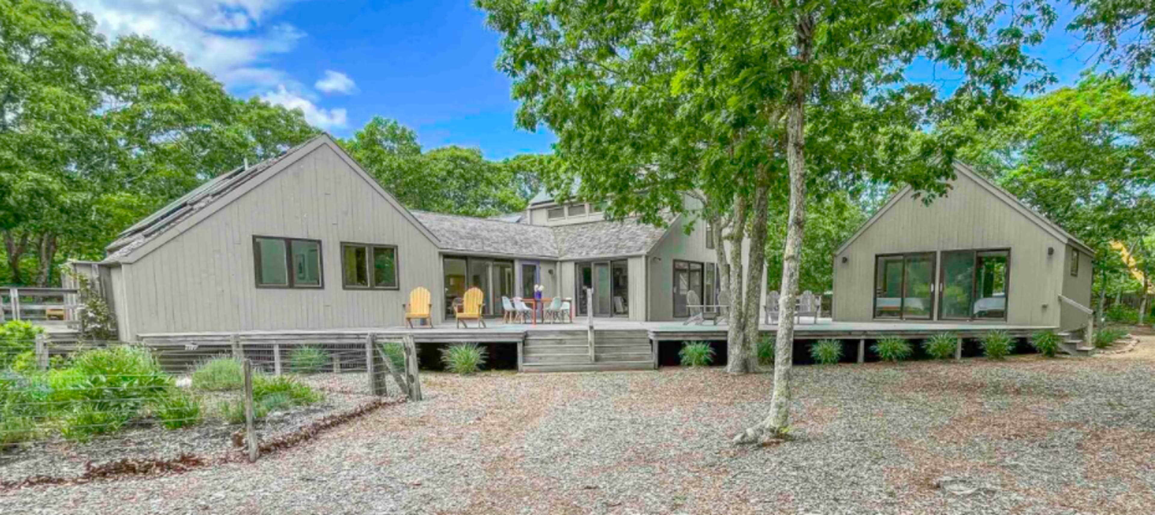 A modern house with multiple gabled roofs is set on a gravel path surrounded by trees.