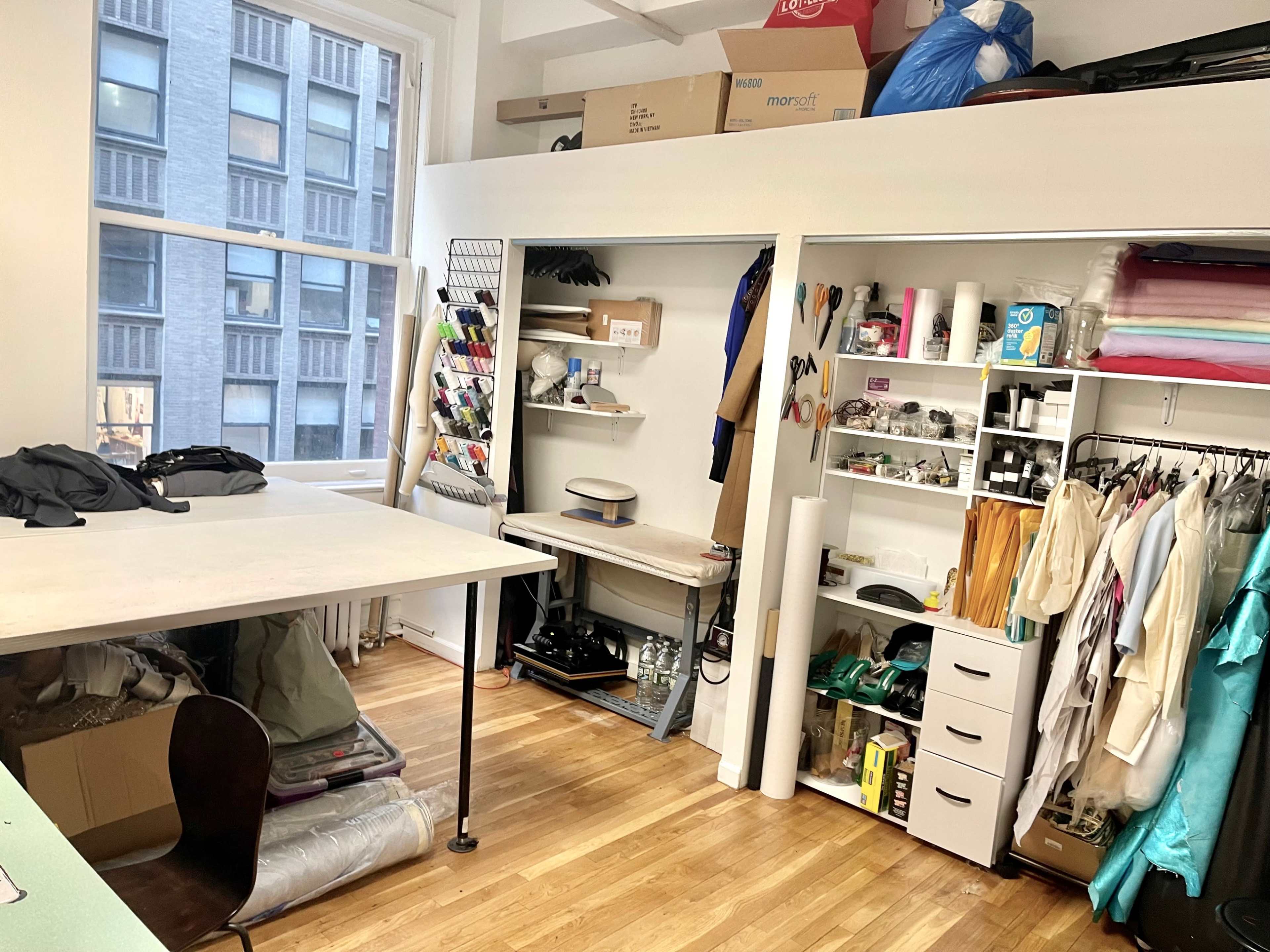 The image shows a cluttered room with a workspace table on the left, shelving units filled with various materials and supplies on the right, and large windows allowing natural light to enter.