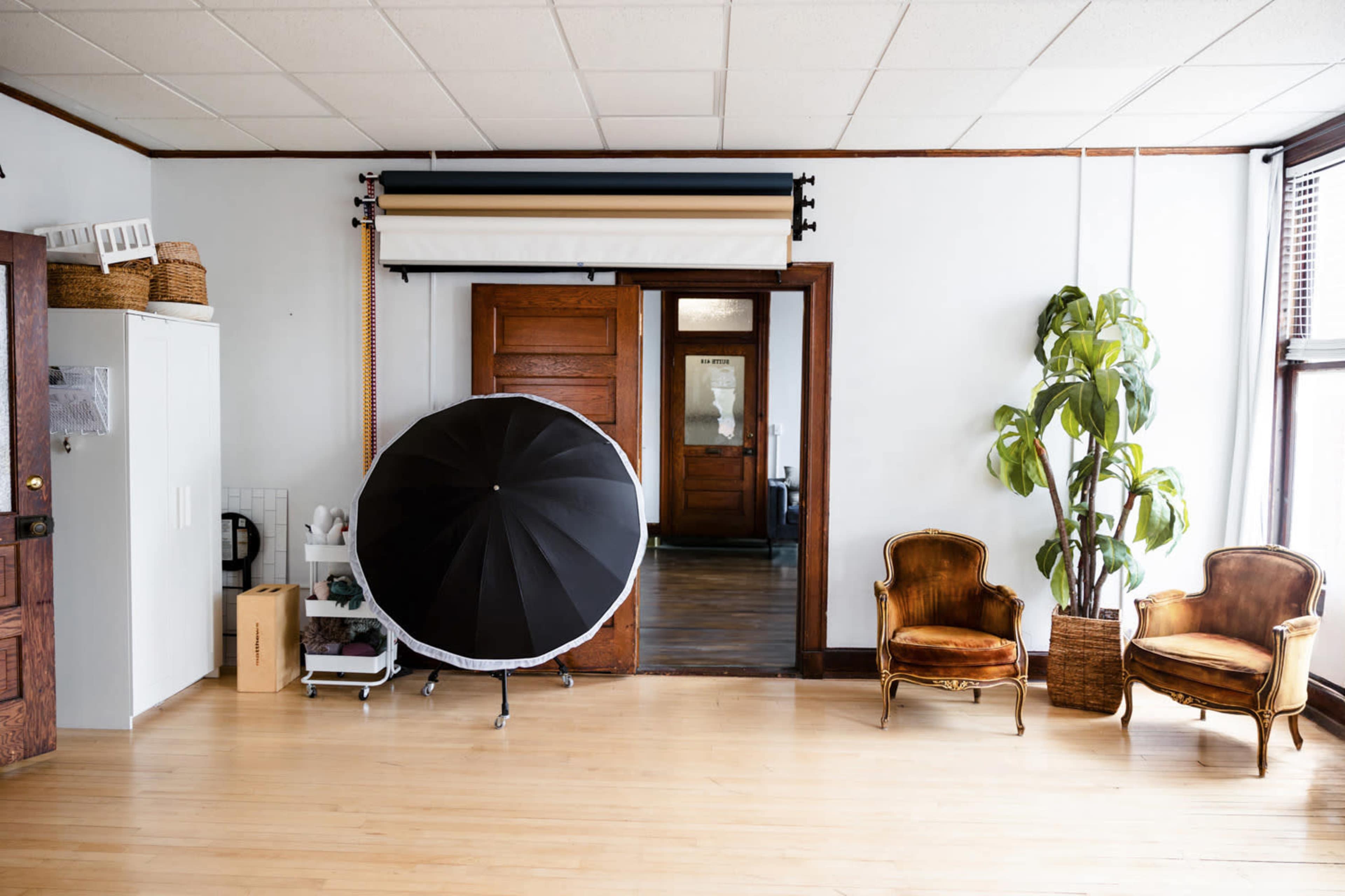 A spacious room with wooden flooring, two vintage chairs, a large circular reflector on a stand, and a plant in the corner, with doors leading to another area.