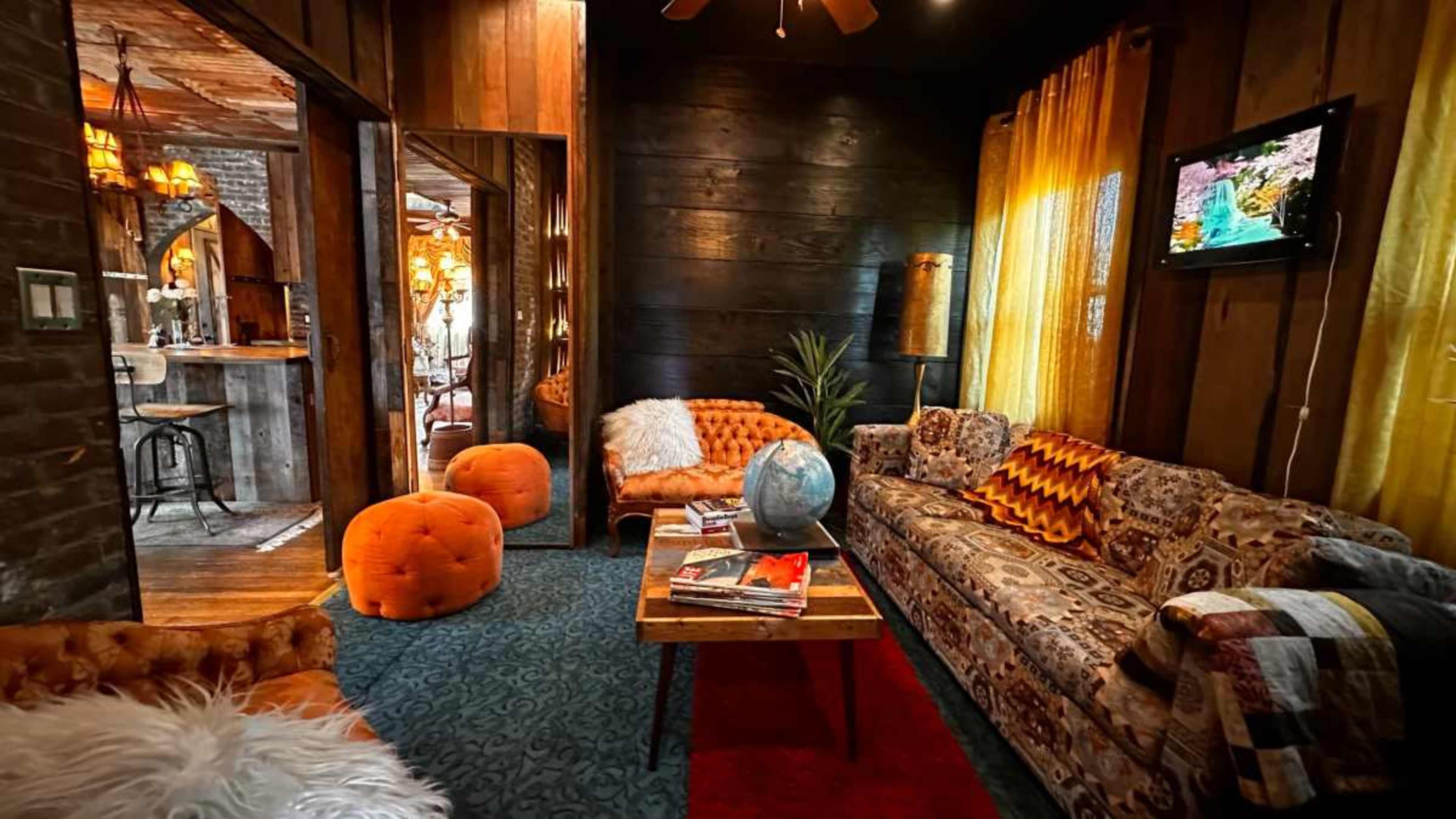 The image shows a cozy living room with vintage furniture, including a colorful patterned sofa and orange poufs, illuminated by warm lighting.