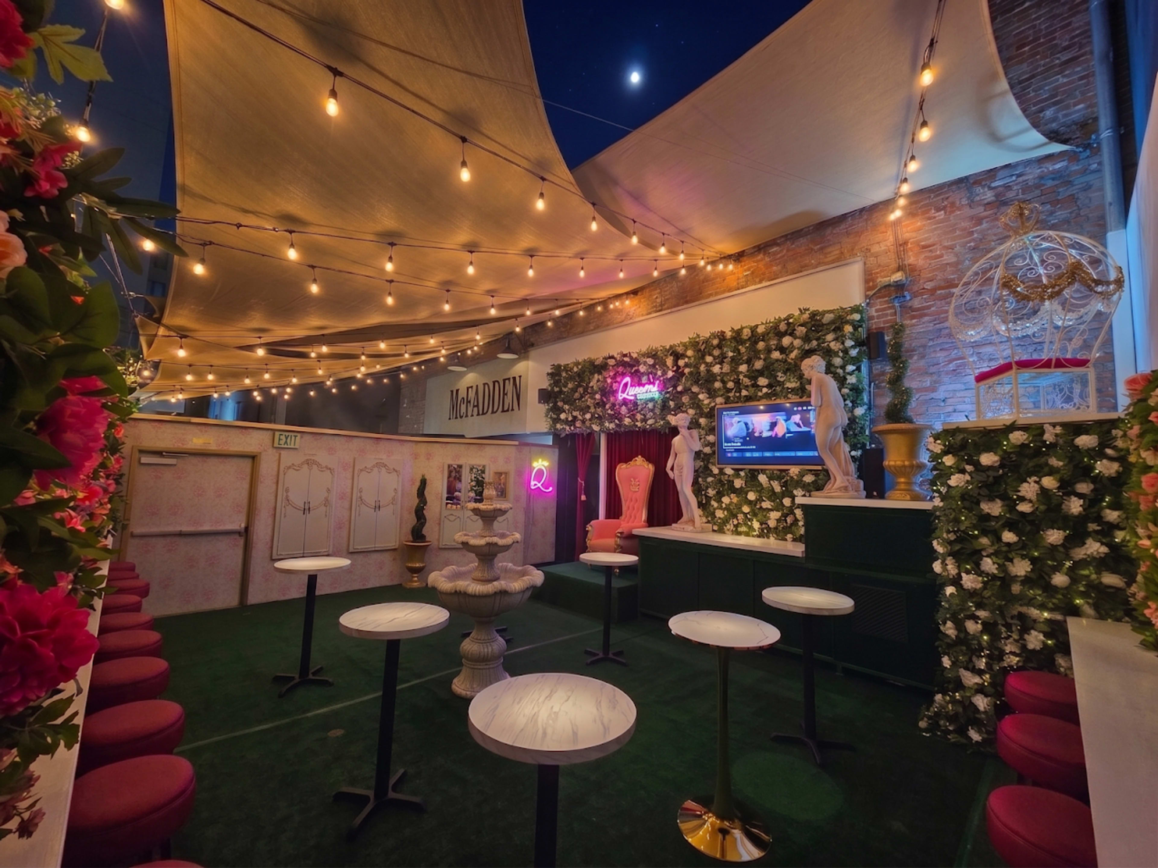 The image shows a decorated outdoor seating area with round tables, a fountain, and a television, surrounded by flower-covered walls and string lights against a night sky.