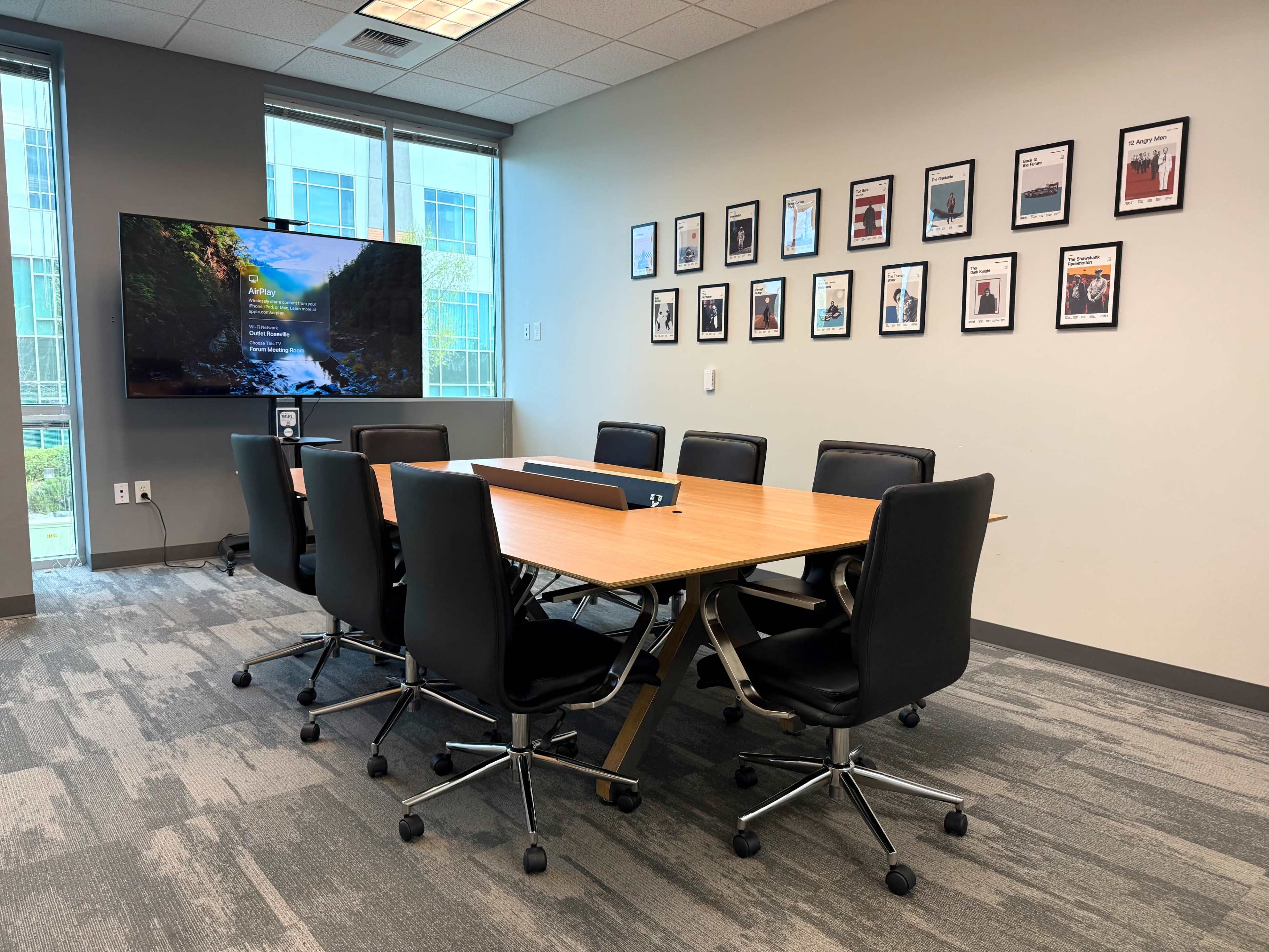 A conference room features a long wooden table surrounded by black chairs, with a large screen mounted on one wall displaying a nature scene, and framed pictures hung on another wall.
