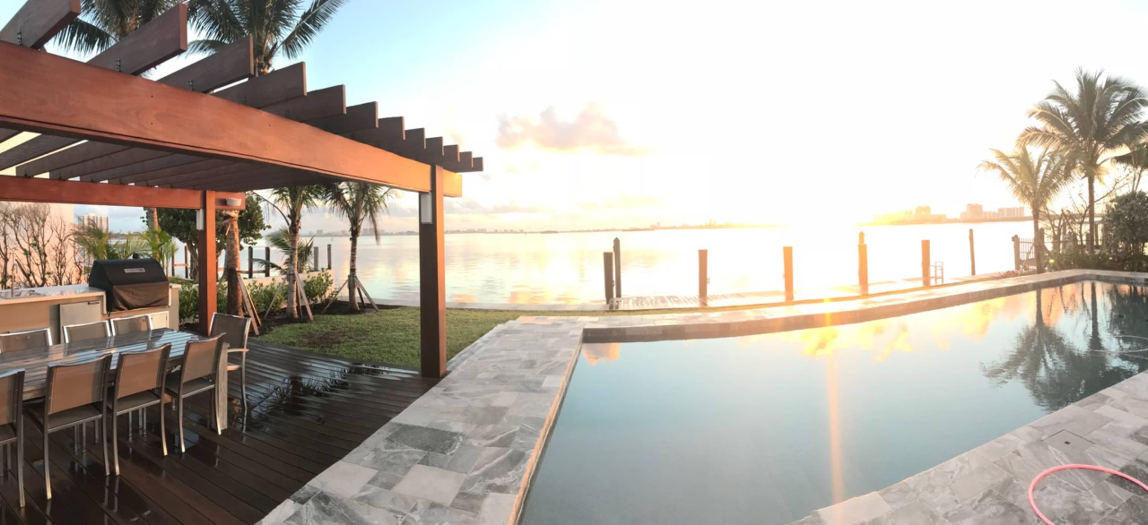 The image depicts a waterfront property with a swimming pool, outdoor dining area, and a clear view of a sunrise over the water.
