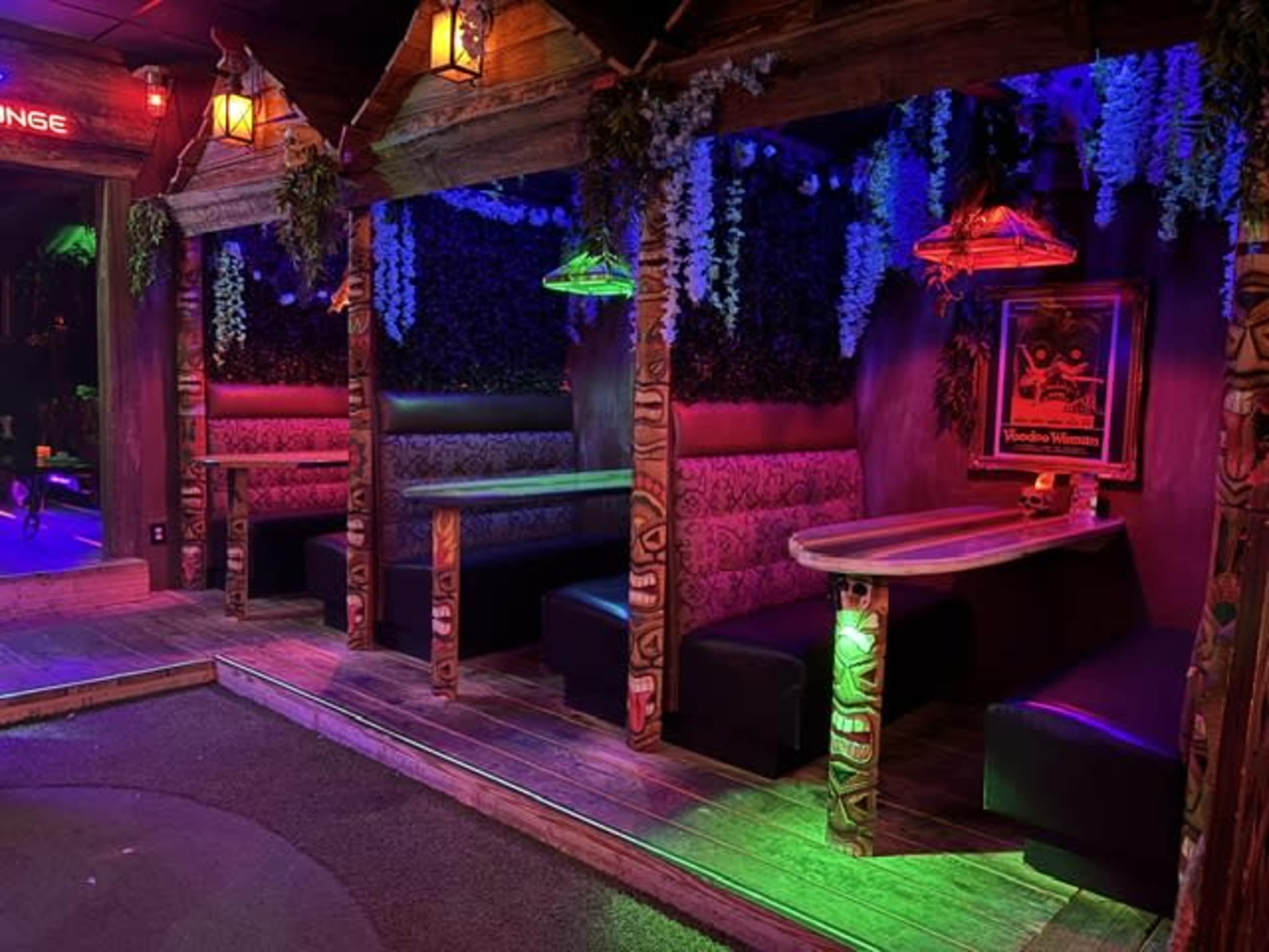 The image features a vibrant lounge area with low, cushioned seating, wooden posts adorned with carvings, and colorful neon lighting creating an inviting atmosphere.