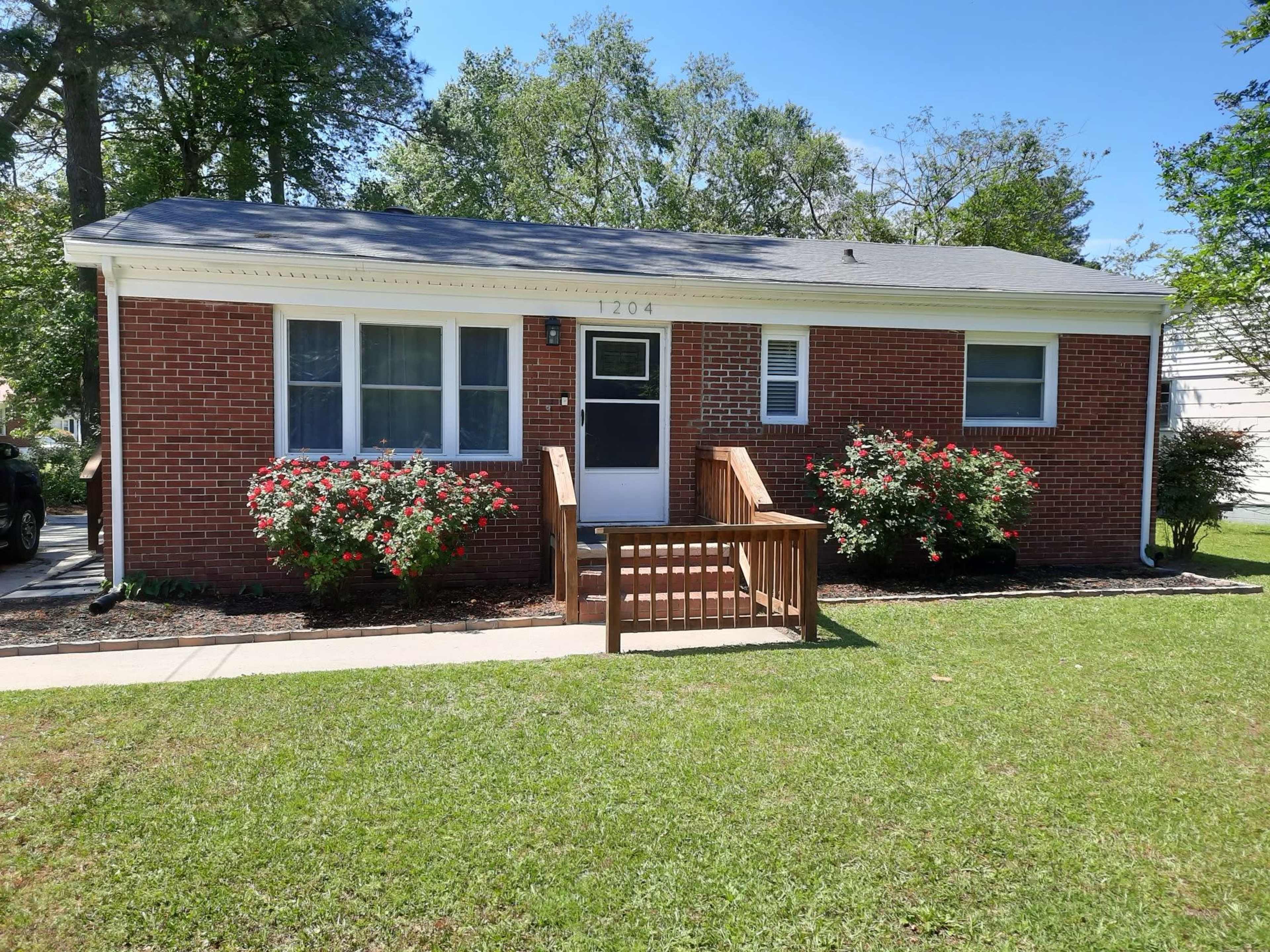 A single-story brick house with a front wooden ramp and flower beds is set on a grassy lawn.