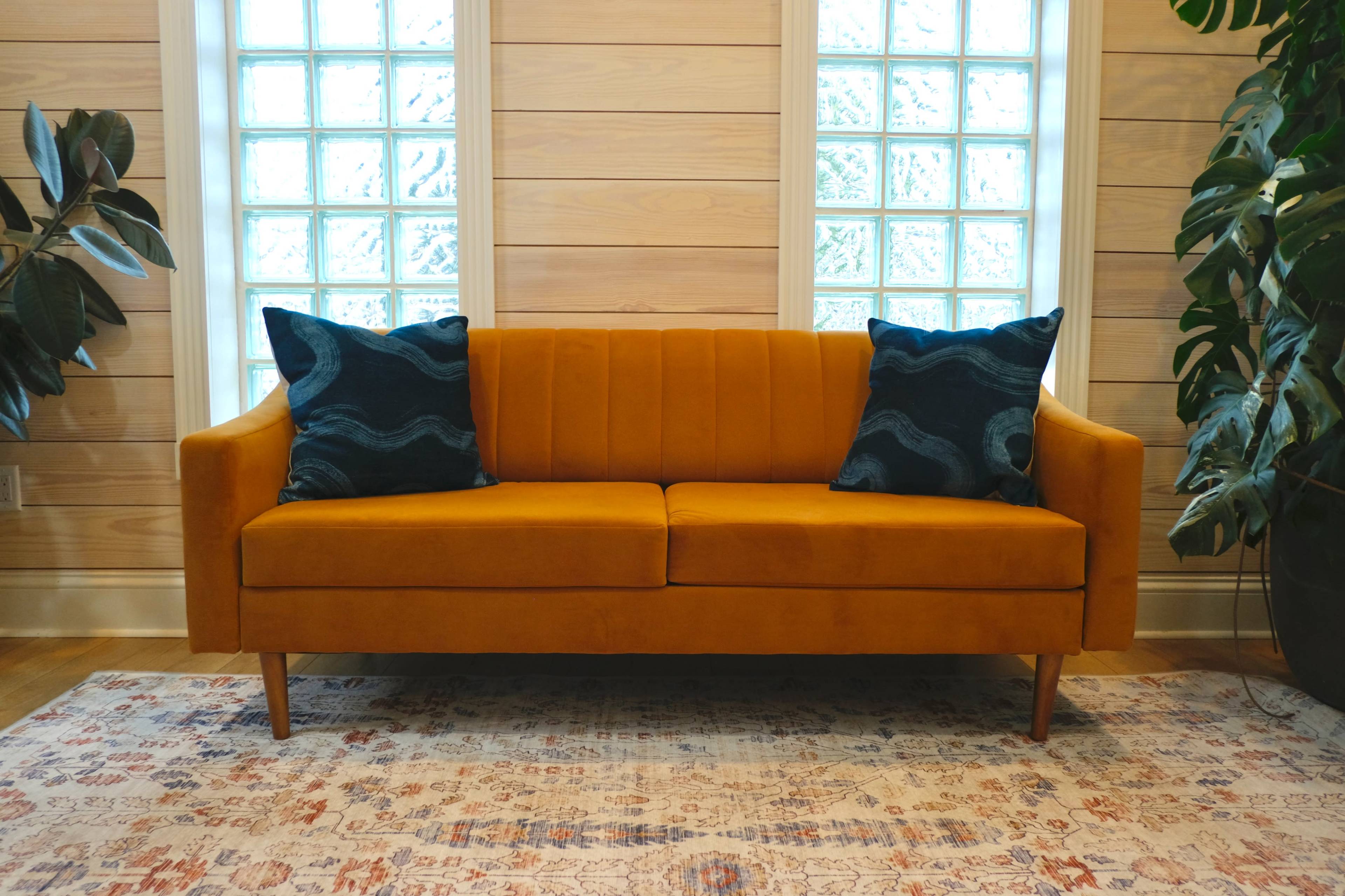 A bright orange couch with two decorative pillows sits against a wooden wall with glass block windows.