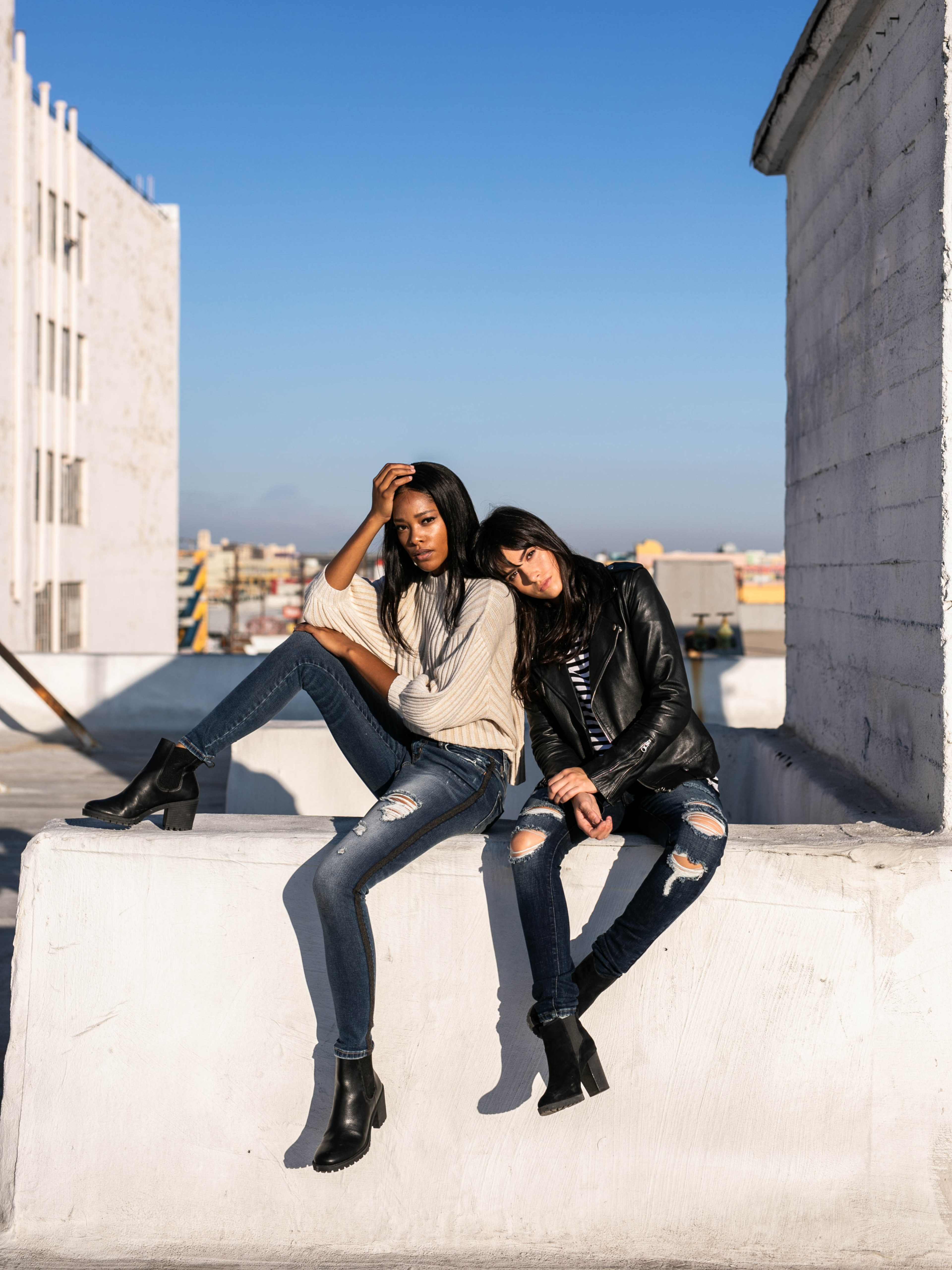 Two women sit on a concrete ledge wearing casual outfits, with one leaning back and the other resting her head on the first's shoulder, against a clear blue sky.