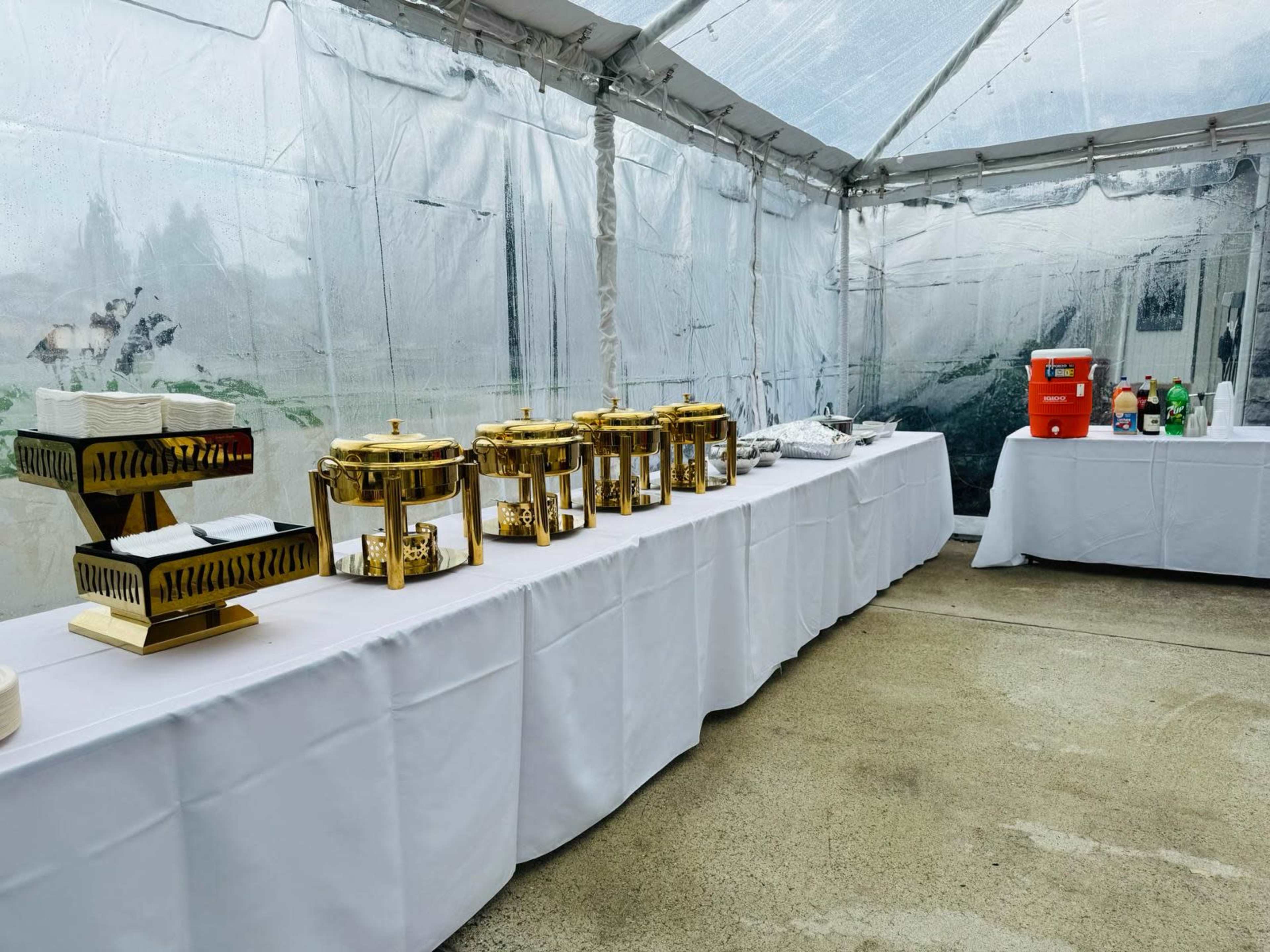A white tent is set up with a long table featuring gold-colored serving dishes and a drink cooler on the side.