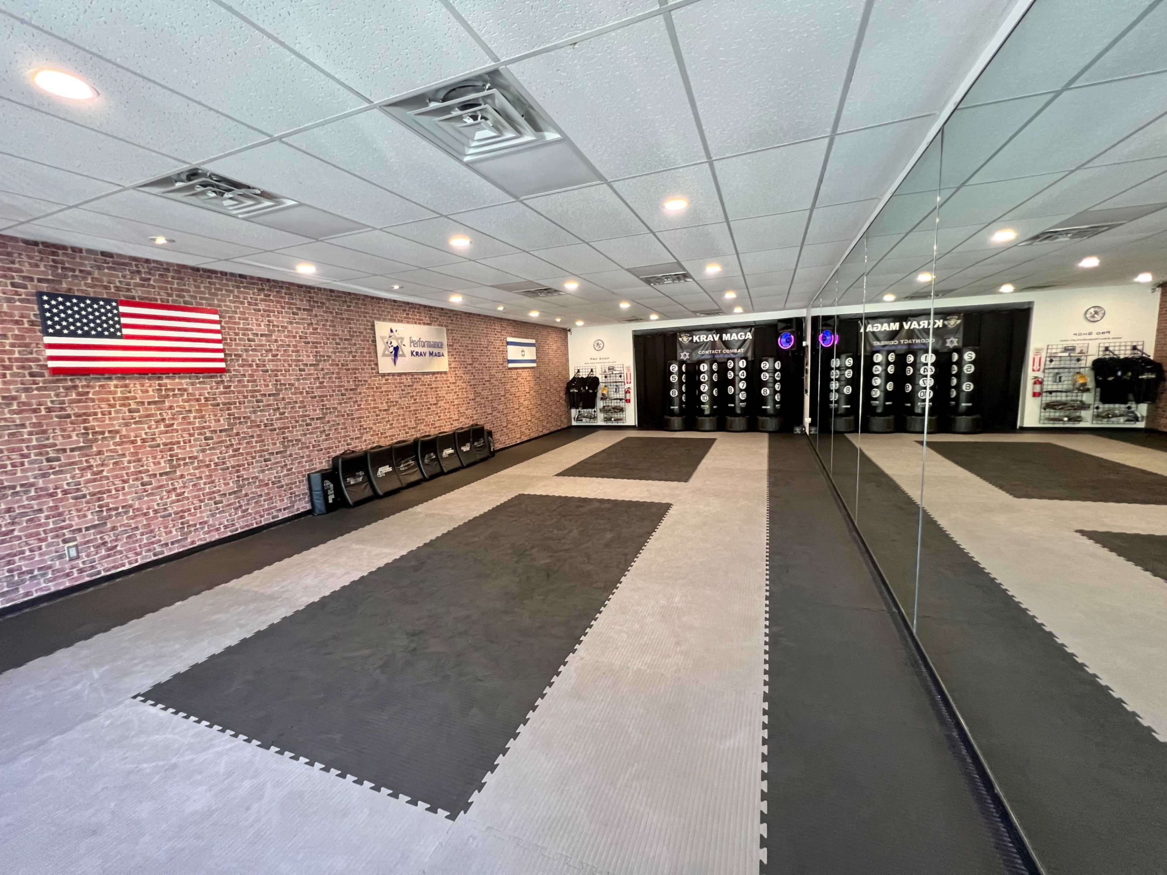 A sparsely decorated martial arts studio featuring a mirrored wall, padded flooring, and an American flag displayed on a red brick wall.