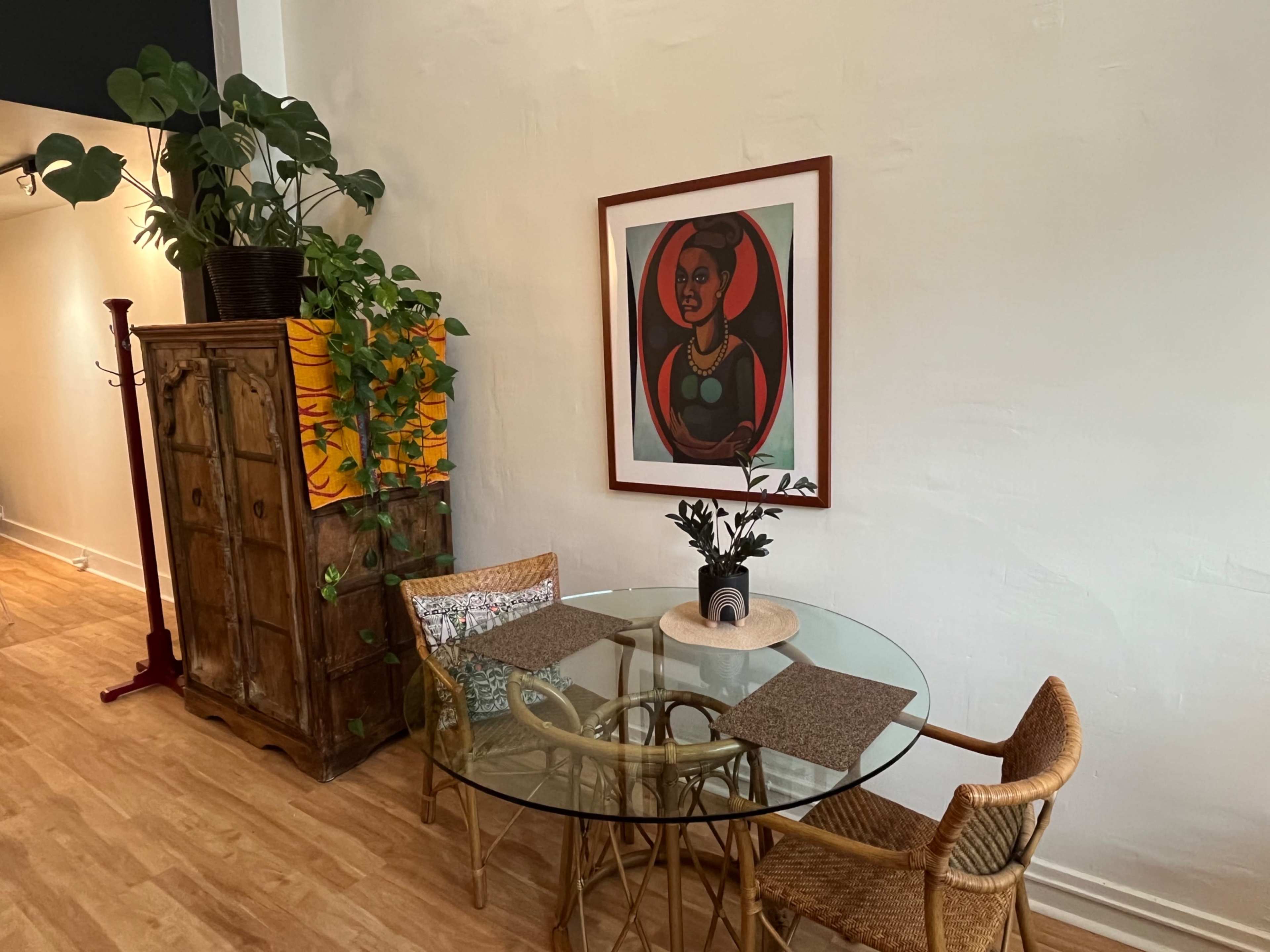 A round glass dining table with woven chairs is positioned near a wooden cabinet and a colorful framed artwork, surrounded by potted plants against a light-colored wall.